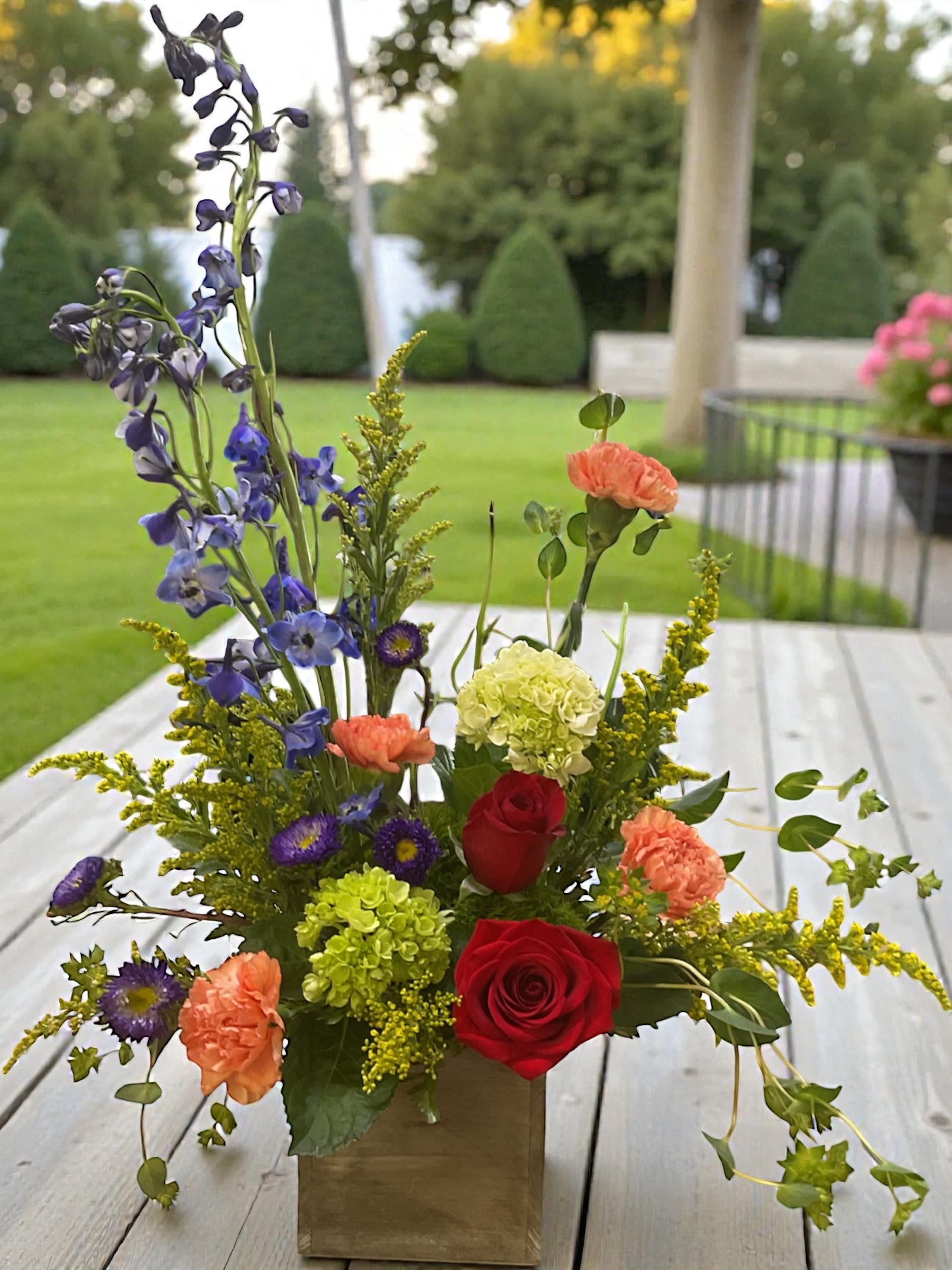 Garden Burst - Bold, vibrant, and absolutely alive with color — Garden Burst is that “walk into a summer garden after the rain” moment in a vase. Electric blue delphinium, fiery red roses, peach carnations, vivid purple asters, bright green hydrangea, and touches of golden solidago explode together in a rustic wooden box for a design that’s fresh, fun, and full of personality.  Send it for birthdays, congratulations, pick-me-ups, thank-yous, or anytime someone needs a bright pop of joy dropped right on their doorstep. This one turns heads — in the best way.  Colors may vary slightly based on seasonal availability. 