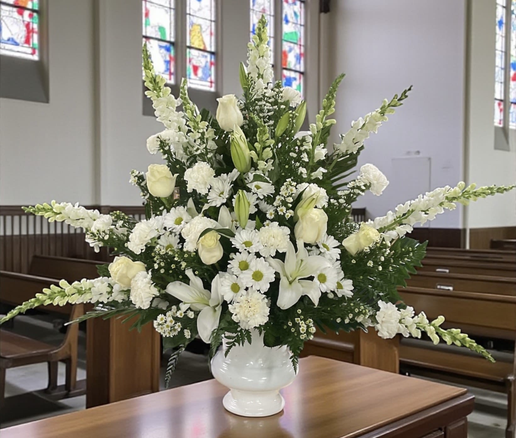 Legacy of Love - Legacy of Love” is a timeless, all-white tribute created to honor a life remembered with dignity and tenderness. Crisp white snapdragons rise with quiet strength, surrounded by pure white lilies, roses, carnations, daisies, and Montecasino for soft texture and peaceful beauty.  Every bloom works together to create a serene, comforting presence — a heartfelt expression of respect, remembrance, and enduring love. Perfect for families who want a classic, elegant sympathy piece that speaks with grace when words are hard to find.