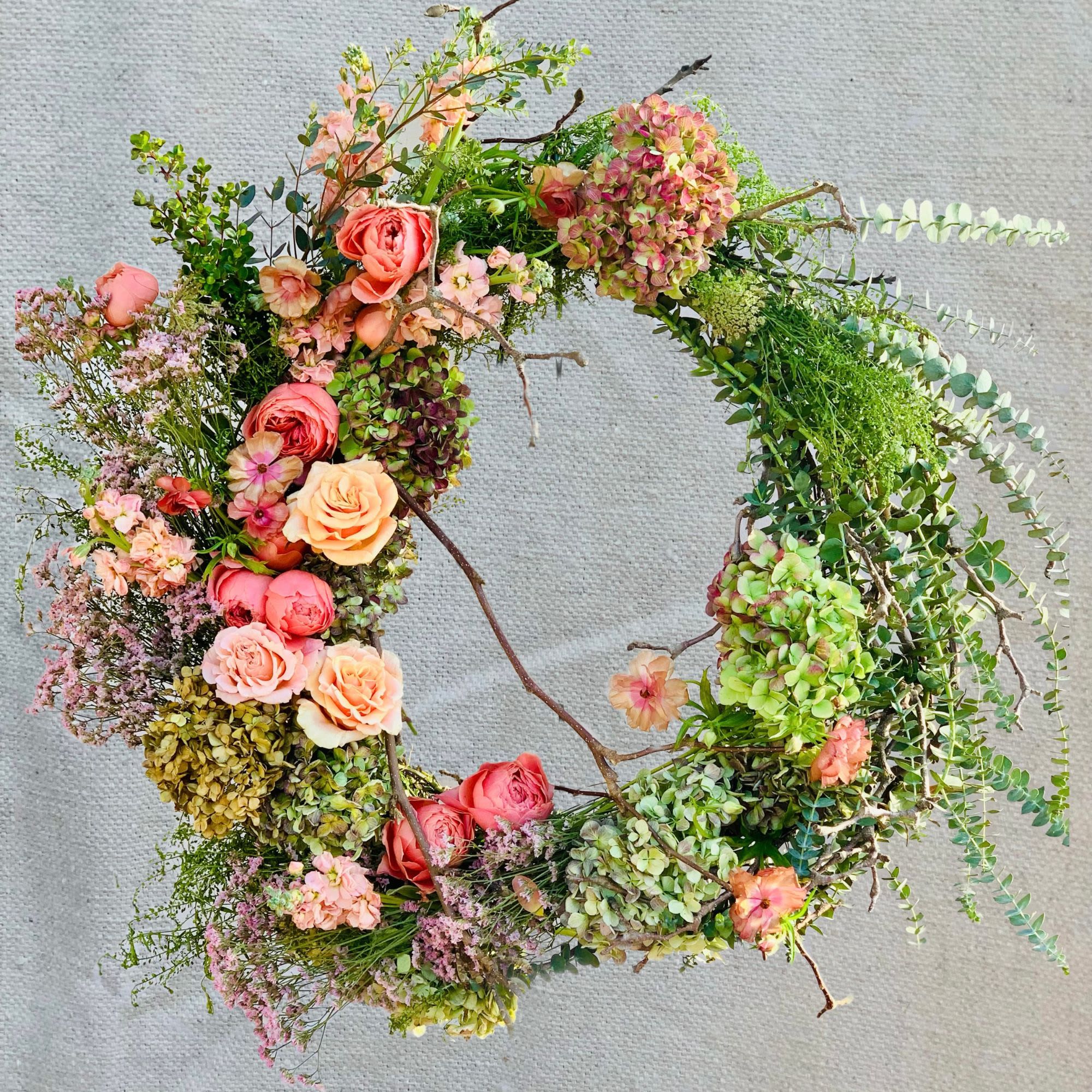 Heavenly Garden - A stunning asymmetrical garden wreath featuring coral and peach garden roses with lush greenery and cascading eucalyptus on a natural grapevine base. This artfully designed tribute combines garden roses, butterfly ranunculus, antique hydrangea, stock, misty blue, natural branches, and abundant eucalyptus, creating a naturally elegant expression of beauty and remembrance with organic, flowing movement.  Specific florals can be included or removed and colored themes are available. Each wreath includes a 5' wire easel and is expertly crafted with the freshest blooms and greenery.