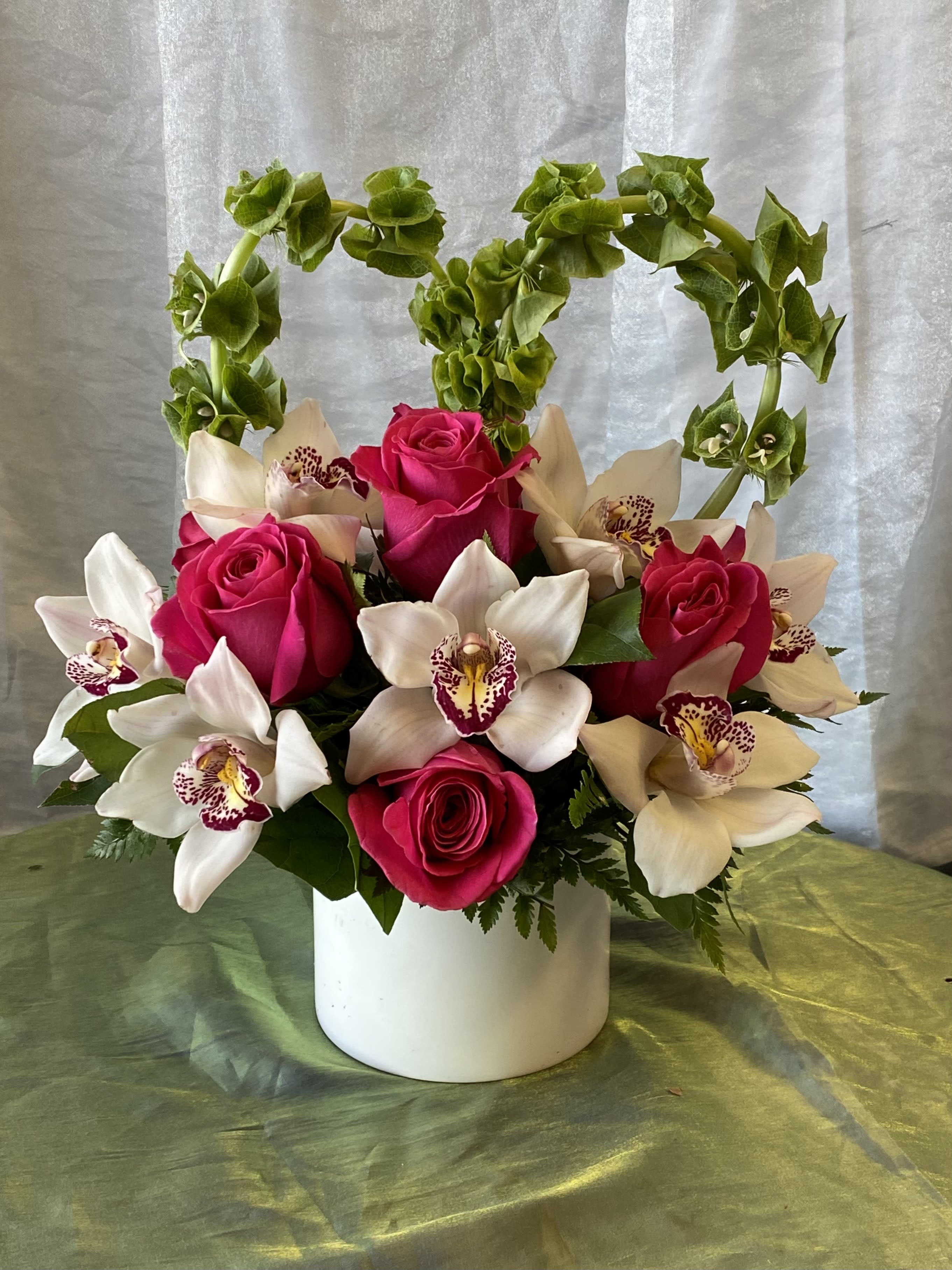 #77 - Mi Chula's Heart - This White Ceramic Container ( will be in BLACK ceramic container)is filled with hot pink roses and white cymbidium orchids. Bells of Ireland are tied together to create a heart shape slightly above the flowers. ***will be in BLACK ceramic container**