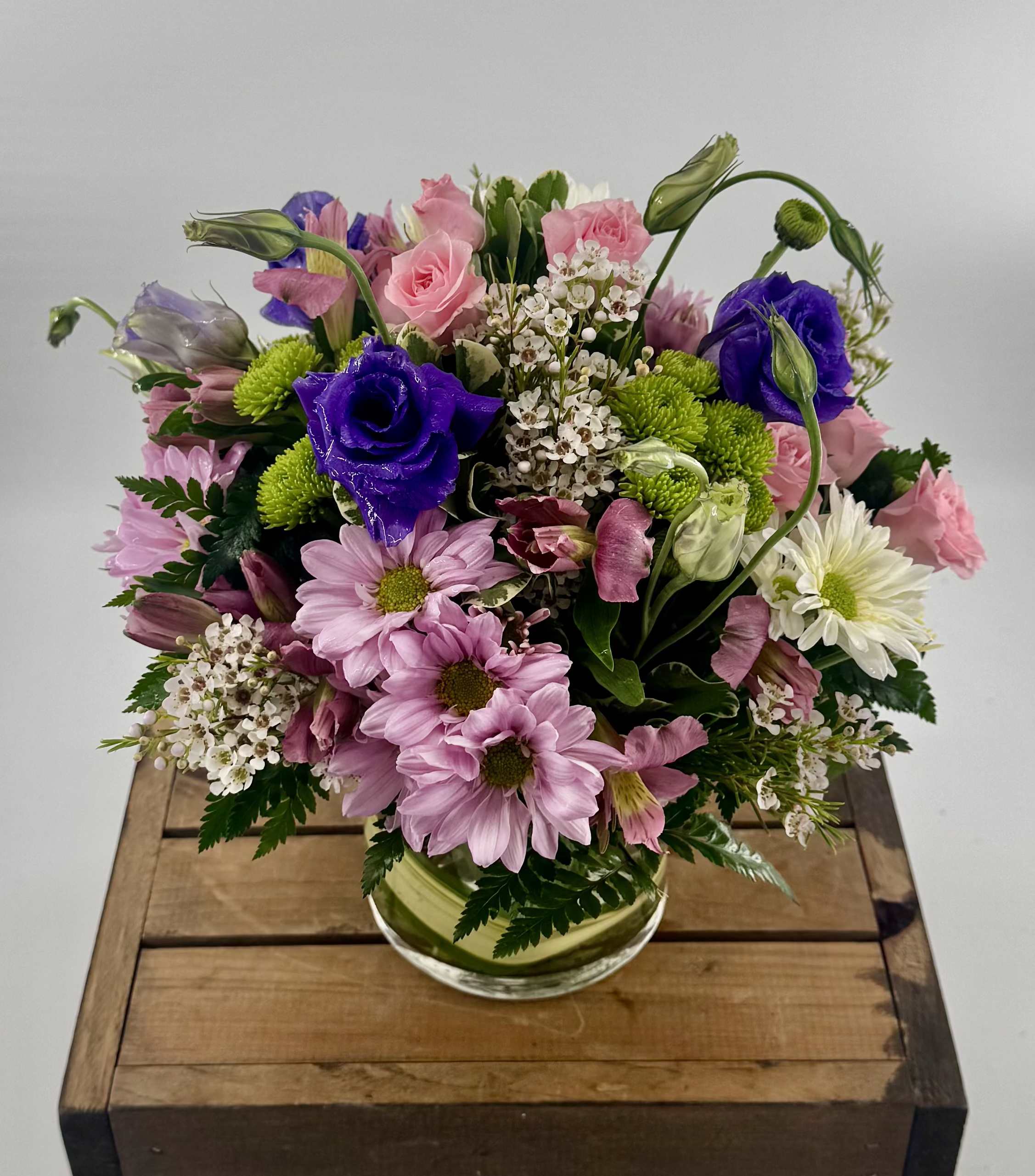 Berry Bliss (BR) - Introducing our Berry Bliss Floral Arrangement, a vibrant and charming display of nature's beauty. This delightful arrangement features a mix of purple lisianthus, green button poms, pink spray roses, white and lavender daisies, pink alstroemeria, waxflower  all lovingly arranged in a clear glass 5"x5" cylinder vase.