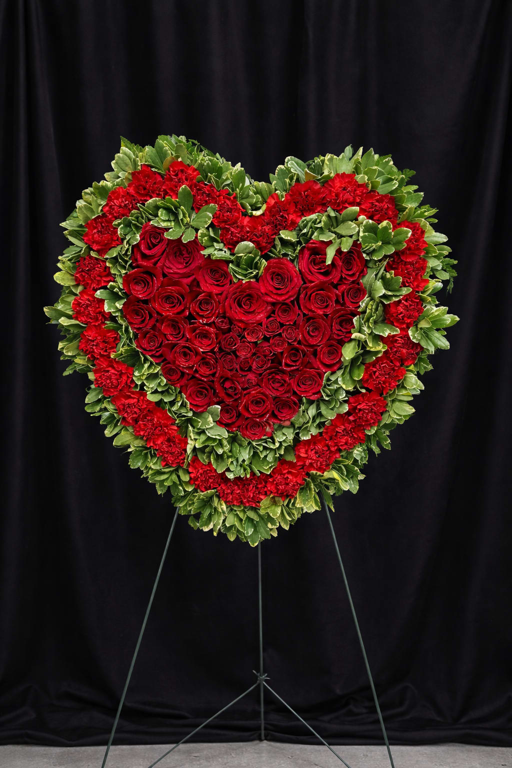 Crimson Devotion - Crimson Devotion is a timeless expression of deep love and remembrance. Designed in a classic heart silhouette, this standing tribute features a lush center of premium red roses surrounded by vibrant red carnations and rich greenery for texture and depth. The layered composition creates dimension while allowing the velvety roses to take center stage.  Displayed on a standing easel, this piece makes a powerful and elegant statement at funeral services, memorials, or celebration-of-life gatherings. Crimson Devotion symbolizes enduring love, respect, and heartfelt remembrance — a beautiful way to honor someone who will forever hold a place in your heart.