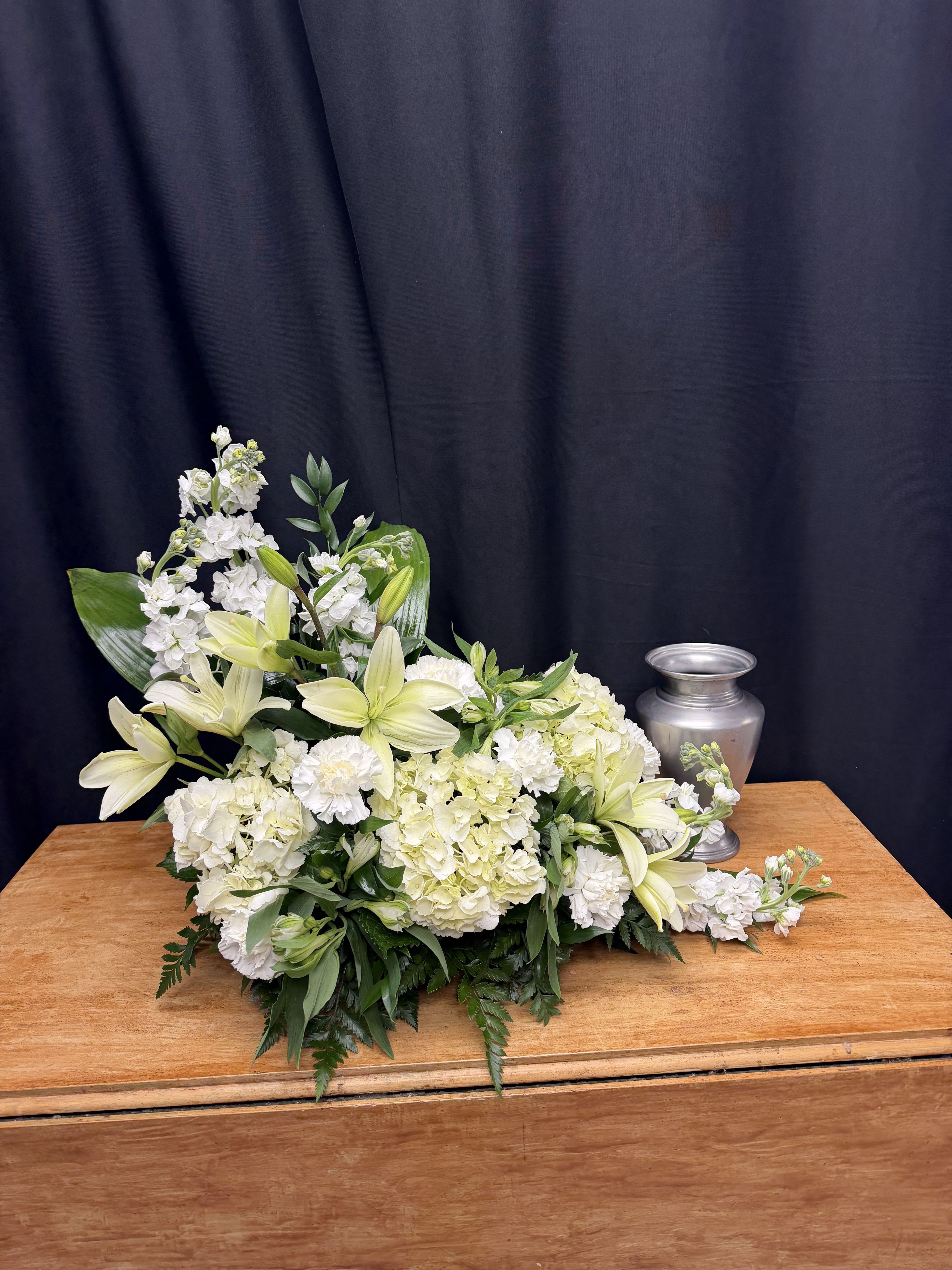 Quiet Grace - Quiet Grace is a refined and elegant cremation tribute designed in a serene palette of soft whites and gentle greens. Featuring graceful lilies, lush white hydrangea, delicate stock, and textured chrysanthemums accented with fresh greenery, this design creates a peaceful and dignified tribute.  The natural, flowing composition offers softness and movement while maintaining a classic silhouette appropriate for traditional services. Quiet Grace symbolizes purity, remembrance, and everlasting peace — a beautiful expression of honor for a life deeply loved and gently remembered.