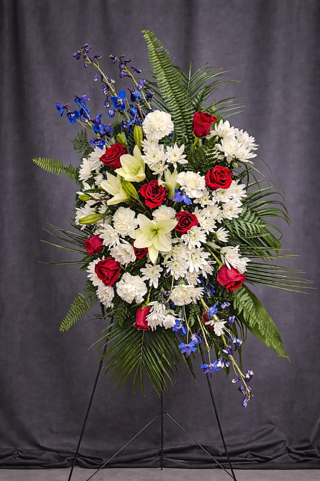 Patriotic Remembrance - Patriotic Remembrance is a bold and dignified standing tribute designed to honor a life of service, strength, and devotion. This striking arrangement features vibrant red roses, crisp white chrysanthemums and carnations, and vivid blue delphinium, thoughtfully arranged with graceful lilies and lush greenery for height and movement.  The classic red, white, and blue palette creates a powerful symbol of love, respect, and national pride. Displayed on a standing easel, Patriotic Remembrance offers a meaningful expression of gratitude and remembrance — a beautiful way to pay tribute to a veteran, public servant, or someone deeply cherished.
