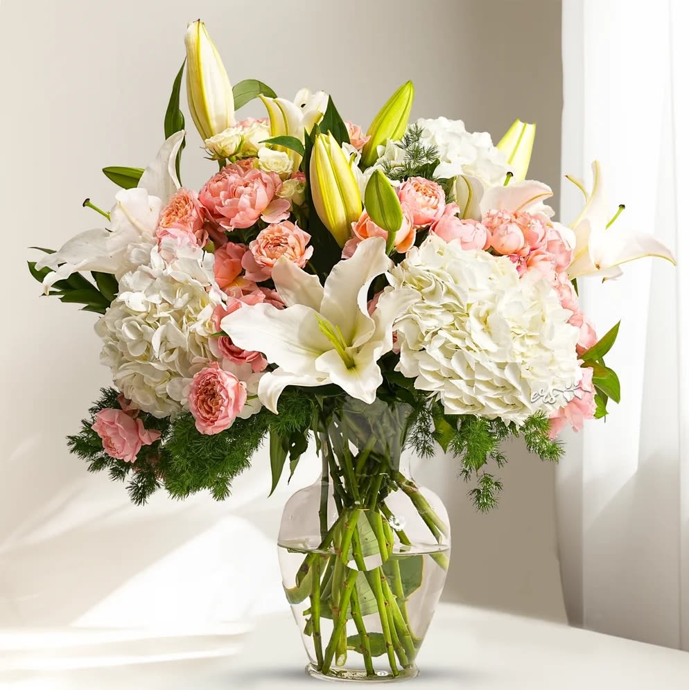 Exquisite Arrangement  - Embrace the serene beauty of our elegant arrangement featuring  white hydrangeas,  spray roses, 5white lilies, and white spray roses, all accented with lush green fillers. Presented in a sleek glass vase, this exquisite ensemble radiates purity and sophistication. Order now to bring a touch of tranquillity and elegance to any space or to delight someone special with this timeless floral display!