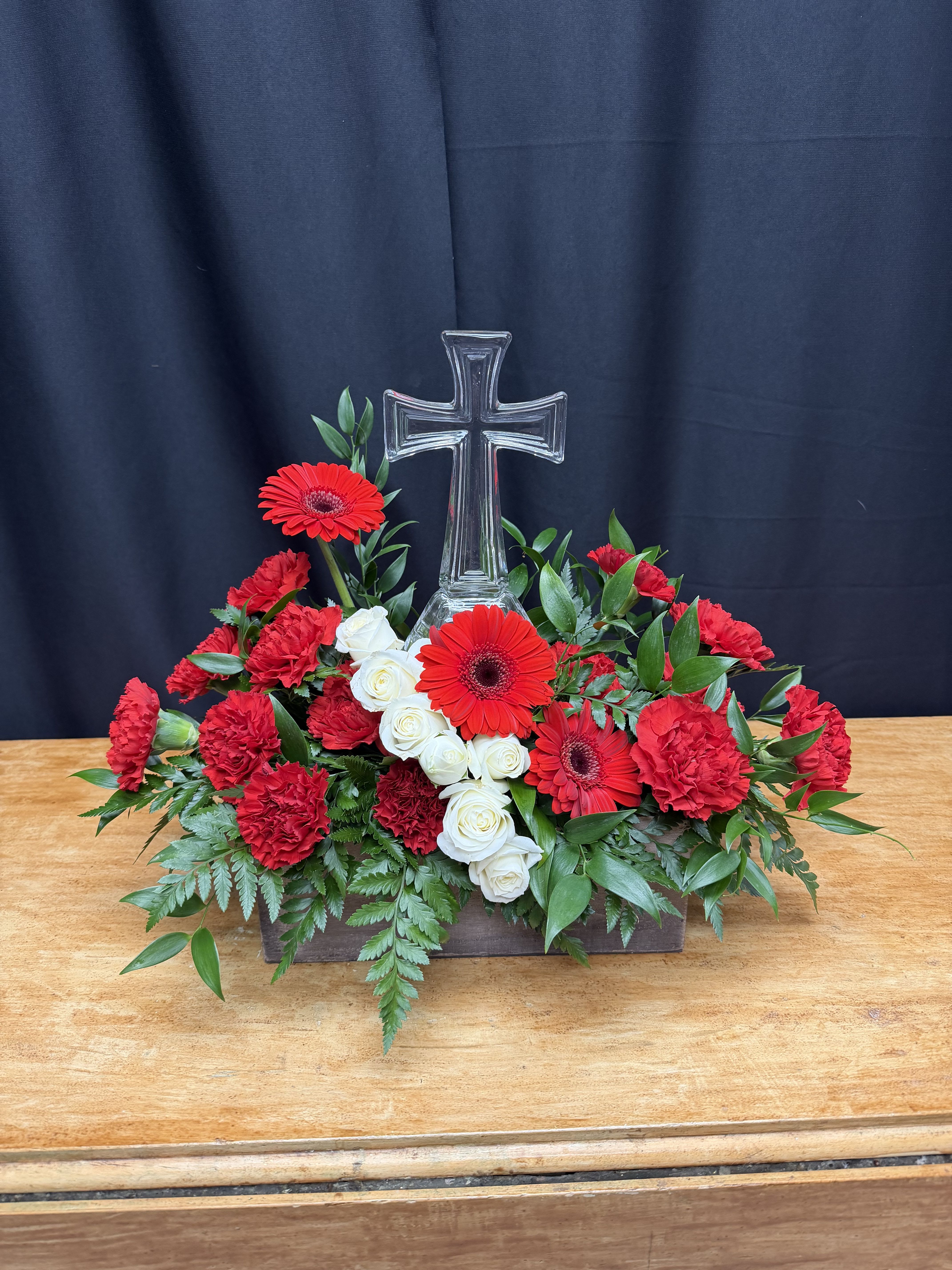 Crimson Faith Tribute - Crimson Faith Tribute is a striking expression of love, devotion, and remembrance. This elegant arrangement features vibrant red carnations and bold red gerbera daisies, beautifully contrasted with classic white roses to symbolize purity, honor, and everlasting peace. Lush greenery cascades naturally from a rustic wooden container, while a clear glass cross rises above the blooms as a powerful symbol of faith and eternal hope.  Designed for memorial services, celebrations of life, or as a heartfelt sympathy gift, this piece offers comfort and a meaningful presence during life’s most tender moments.