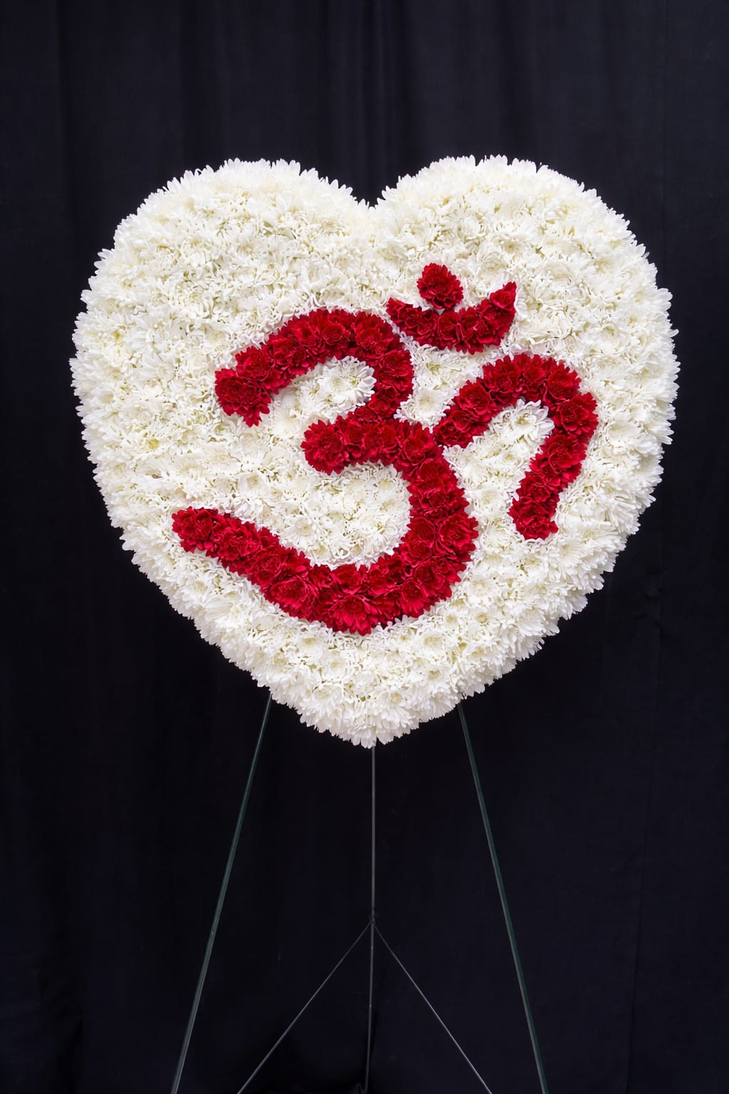 Om Tribute - The Sacred Om Heart is a striking and meaningful standing tribute designed to honor a life rooted in faith, spirituality, and inner peace. This heart-shaped arrangement features a lush foundation of pristine white chrysanthemums, symbolizing purity and eternal love. At its center, the sacred Om symbol is beautifully crafted in vibrant red carnations, representing strength, devotion, and the divine connection of all living things.  Displayed on a standing easel, this distinctive design offers a powerful expression of remembrance for those who embraced yoga, meditation, or Hindu spiritual traditions. The bold contrast of red and white creates a serene yet impactful presence at memorial services or celebrations of life, honoring a soul whose spirit resonates beyond this world.