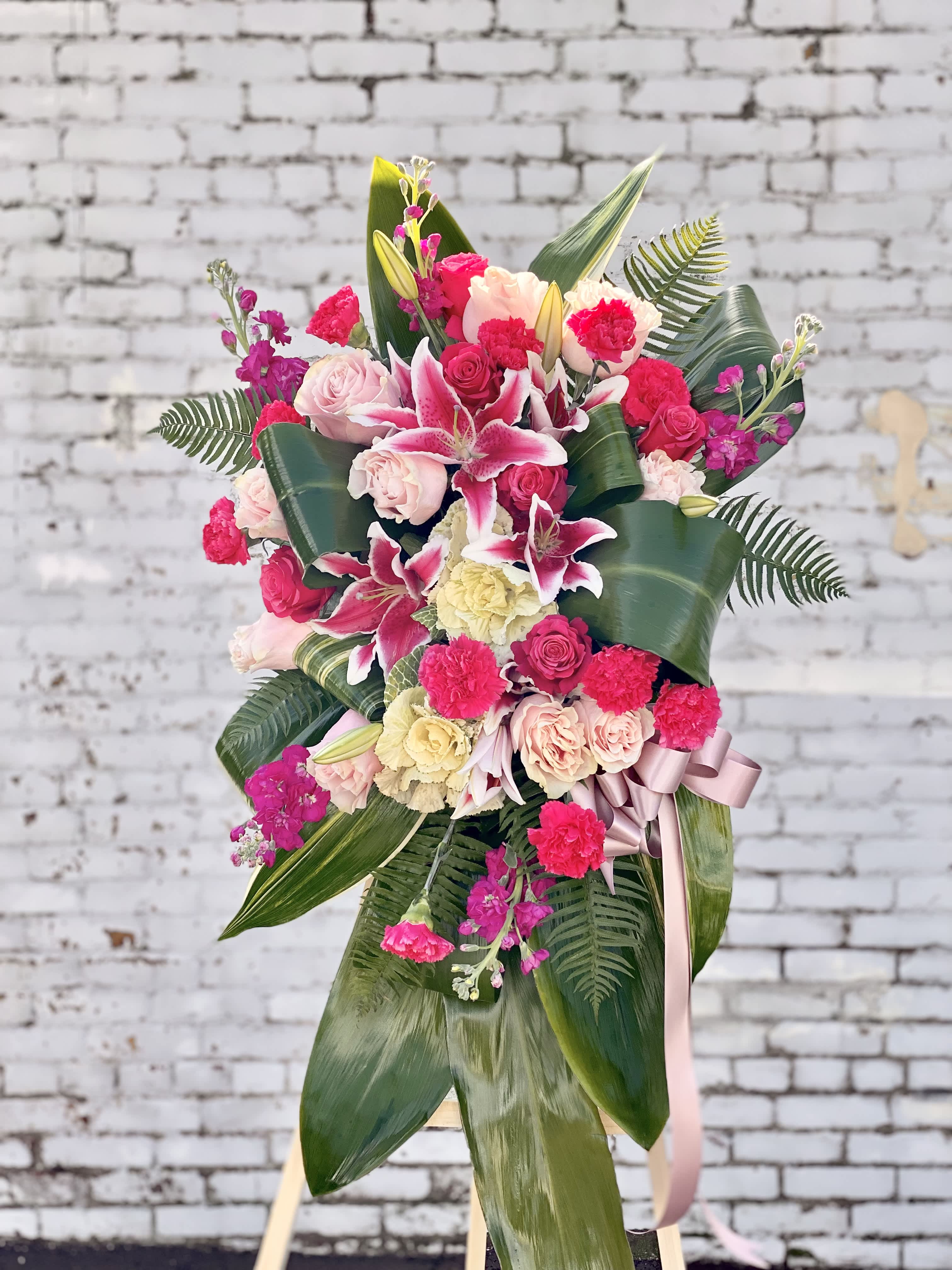 Gently Into Ever After - Bright, fresh, beautiful and just right for an elegant expression of heartfelt sorrow and devastating loss. This classic standing arrangement is constructed by a local FTD artisan florist of hand-selected pink blossoms ranging from pale pastels to deep blushing pink. It includes roses, carnations, stock, cushion pompons, Asiatic and Stargazer lilies and hydrangea beautifully set among a field of lush contrasting greens like aspidistra and ivy vines. It comes with a wooden easel for flexible display options and makes an impressive memorable tribute at a wake, funeral or graveside service.