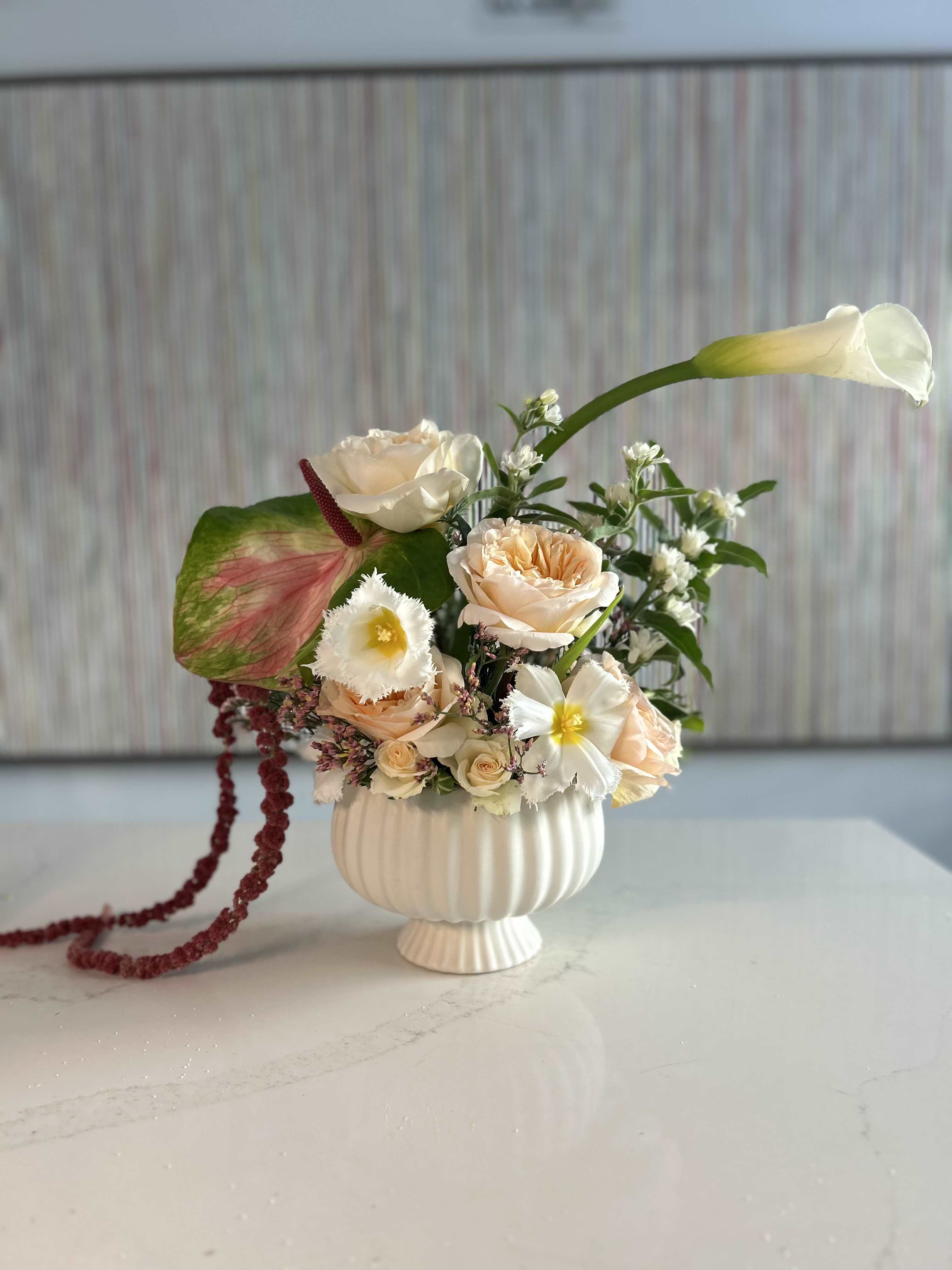 Ivory Poème - Large Calla Lily is the star of the show, supported by gorgeous garden roses, amaranthus, anthurium and fringed tulips  Set in a delightful ceramic compote that is both elegant and playful This arrangement will bring joy and delight to the receiver!