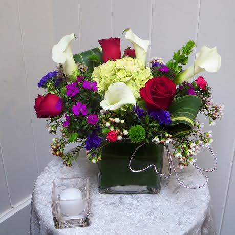 Judith - A funky clear glass cube lined with a ti leaf has hydrangea, roses and mini calla lilies, and a mix of bright filler flowers.
