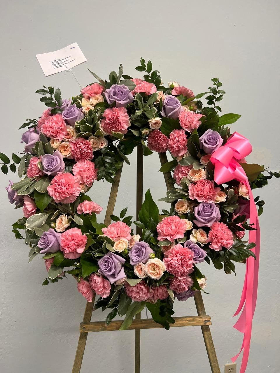 Pink Floral Wreath 18" - A tender expression of love and remembrance, this wreath features a soft and elegant blend of pink carnations, lavender roses, and blush spray roses, accented with lush greenery and finished with a satin pink bow. Its soothing color palette conveys comfort and heartfelt sympathy, making it a beautiful tribute for memorial services or celebration of life ceremonies.