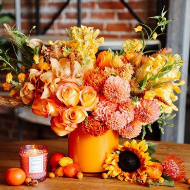 Orange and yellow floral arrangement in a vase with a sunflower and candle nearby