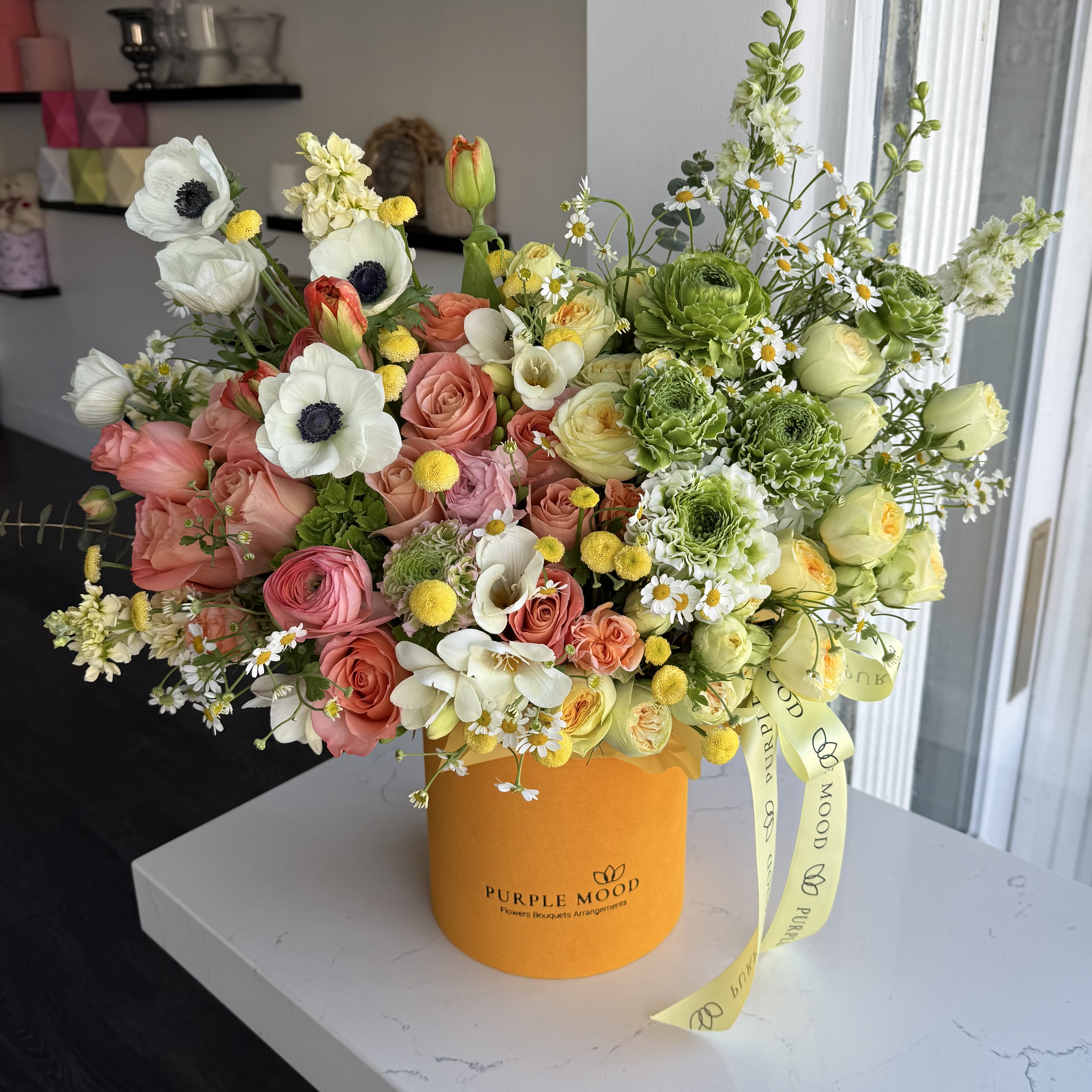 Sunny Meadow Box - A vibrant floral meadow in a box! Sunny Meadow Box features fresh greens, soft peach roses, delicate anemones, and cheerful yellow accents, creating a lush and lively arrangement. Inspired by a sunlit meadow, this design brings warmth, joy, and natural beauty to any space.
