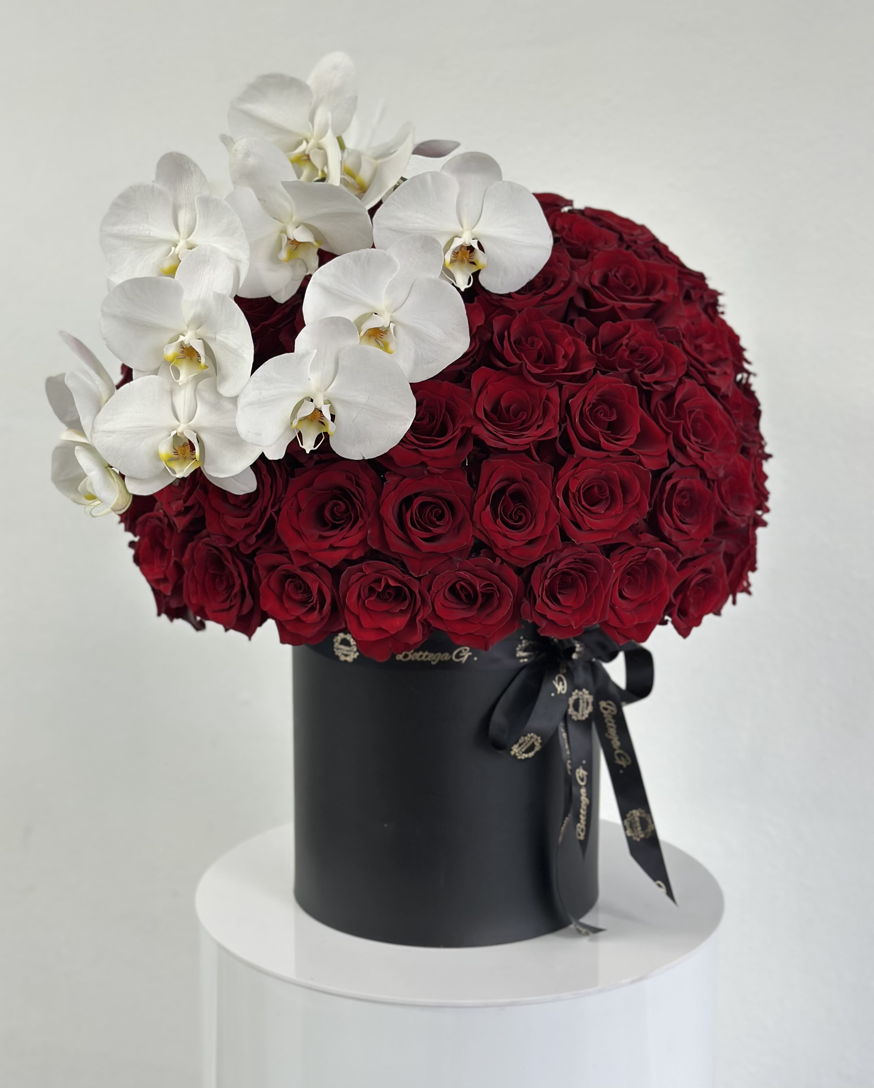 Orchid &amp; Rose Hat Box - A luxurious floral arrangement that combines bold beauty with delicate grace. Inside a sleek, round hat box, vibrant red roses radiate passion and love, while white orchids delicately crown the design, adding a touch of elegance and purity. This stunning combination of flowers is complemented by lush greenery, creating a balanced and visually striking arrangement. Perfect for any occasion that celebrates love, admiration, or remembrance, this hat box arrangement brings a refined beauty and lasting impression.