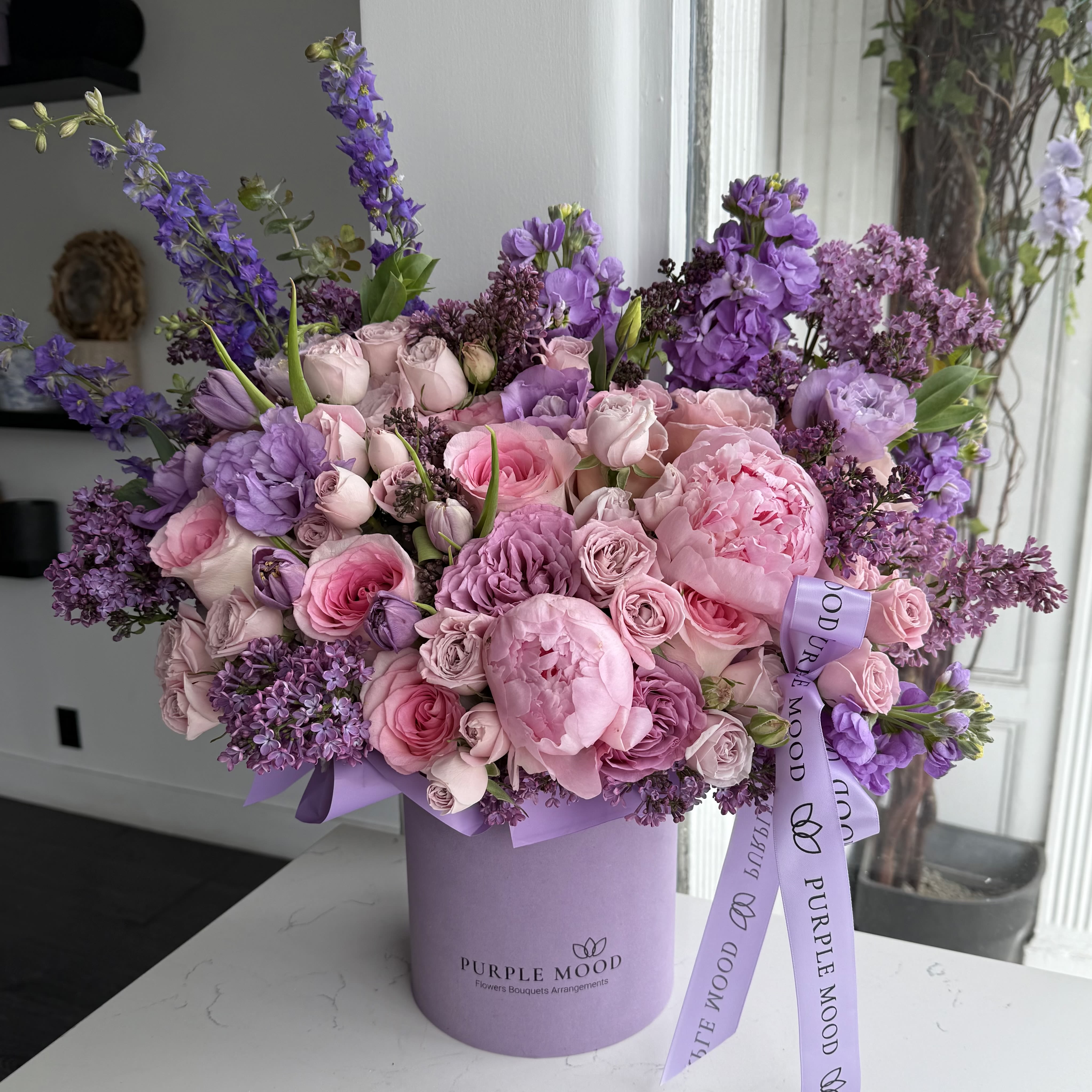 Double Lilac - A romantic blend of soft blush and elegant lavender tones, this lush arrangement is designed in our signature Purple Mood hat box. Featuring premium pink peonies, garden roses, spray roses, and seasonal lilac accents, beautifully complemented with fresh purple blooms for texture and height.  Full, airy, and feminine — this arrangement makes a perfect gift for birthdays, anniversaries, new baby celebrations, or simply to brighten someone’s day.  Designed in our keepsake lavender box with Purple Mood ribbon.  Flower varieties may vary slightly based on seasonal availability while maintaining the overall look and color palette.