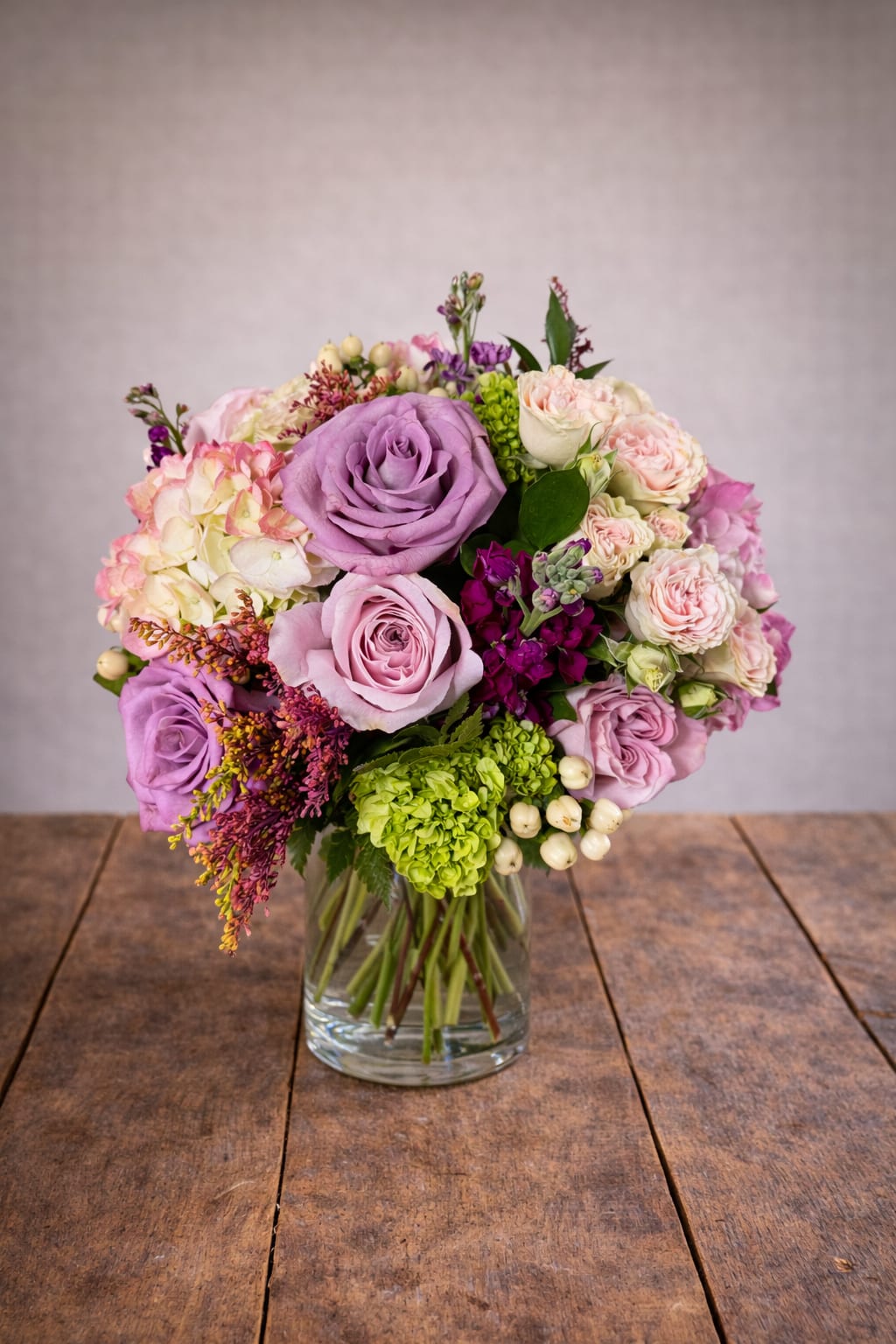 Suzi's Secret Rose Garden - This elegant glass vase is filled with a stunning arrangement of vibrant purple and pink flowers, creating a beautiful and eye-catching centerpiece for any room. The combination of colors and textures adds a touch of elegance and sophistication to any space, making it the perfect addition to your home decor. Whether displayed on a dining table, mantelpiece, or sideboard, this vase of flowers is sure to brighten up any room and bring a sense of natural beauty indoors. 