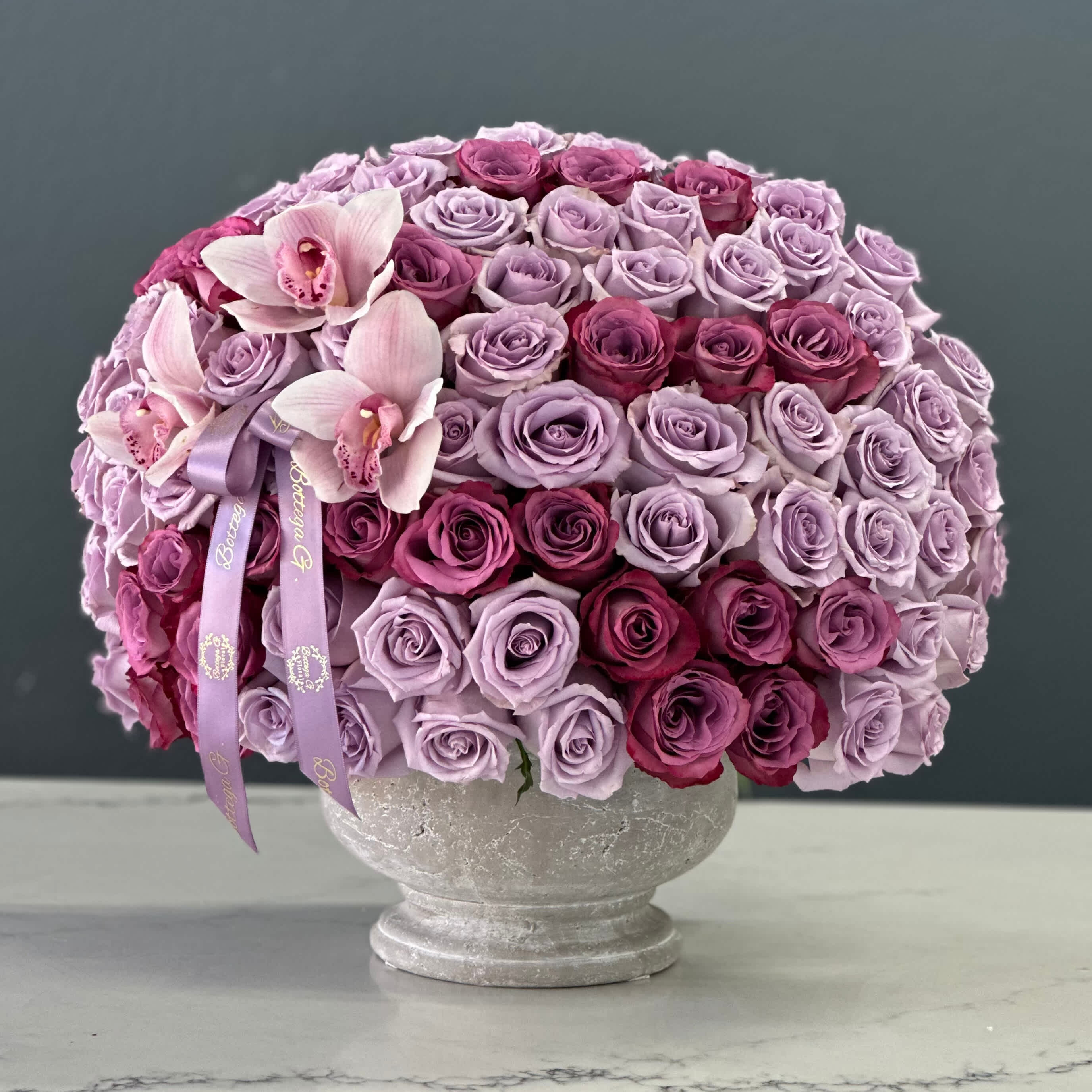 Plum Paradise - Description:  In this exquisite Mother's Day arrangement, we've curated a symphony of nature's finest blooms, marrying the regal allure of purple roses with the ethereal charm of orchids. Each element harmonizes to create a visual masterpiece that embodies love, appreciation, and gratitude.  Flowers:  Purple Roses: Symbolizing admiration, enchantment, and everlasting love, the deep hue of purple roses adds a touch of royalty to the arrangement. Their velvety petals unfurl gracefully, exuding sophistication and warmth.  Orchids: Elegant and enchanting, orchids symbolize rare beauty, refinement, and delicate grace. Their intricate blooms, in shades ranging from soft lavender to rich magenta, create a mesmerizing focal point, captivating the beholder with their enchanting allure.  Vase:  Crafted from durable yet stylish cement, the vase serves as a contemporary canvas, perfectly complementing the organic allure of the flowers. Its rugged texture and neutral tones provide a striking contrast to the softness of the blooms, enhancing their beauty while adding a touch of modern sophistication.