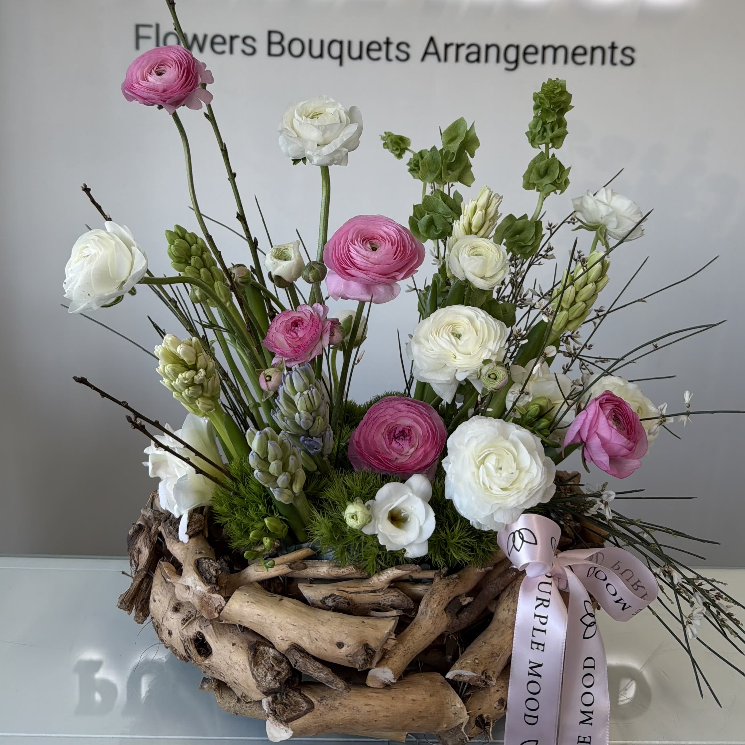 Blush Meadow - A charming garden-style arrangement inspired by a blooming spring meadow. Featuring soft pink and crisp white ranunculus, delicate seasonal blooms, fresh greenery, and natural textural accents, this design is arranged in a rustic driftwood-style base for an organic, airy look.  Light, whimsical, and full of movement, Blush Meadow is perfect for birthdays, thank you gifts, new baby celebrations, or simply bringing a touch of spring indoors.  Each arrangement is uniquely handcrafted. Flower varieties may vary slightly based on seasonal availability while maintaining the overall color palette and style.