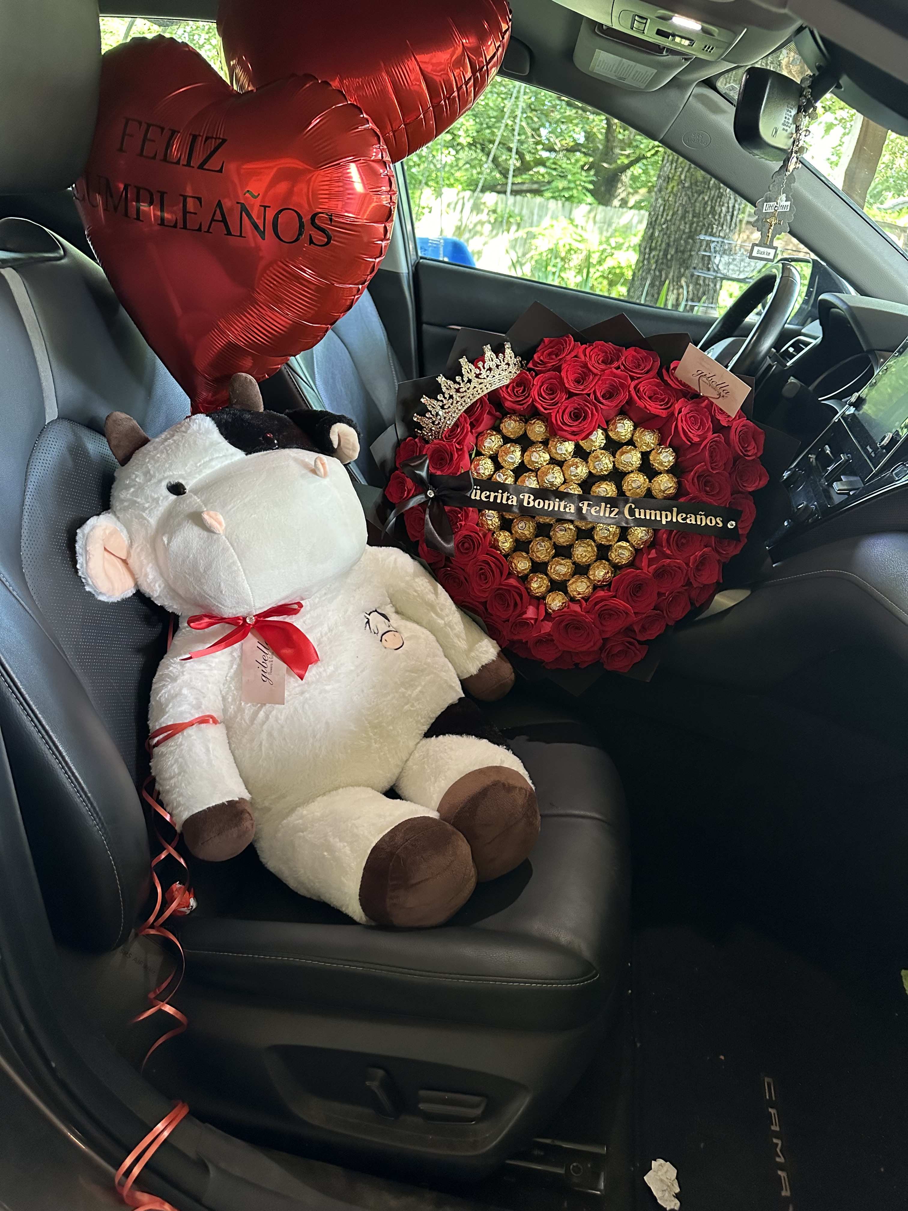 Red heart balloons, a plush cow, and a rose-and-chocolate gift arrangement in a car seat