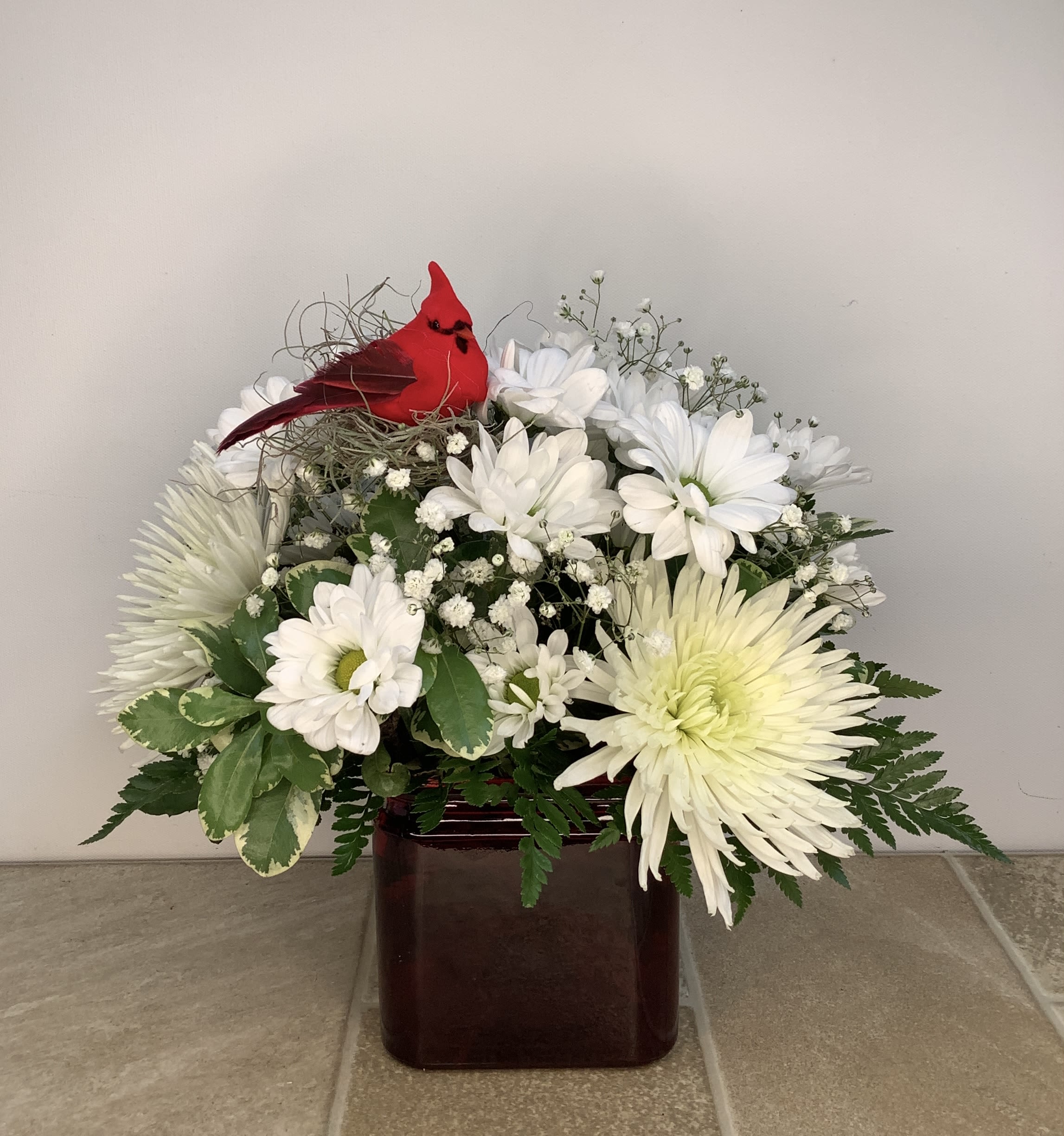Cardinal's Embrace - This elegant arrangement of lovely white flowers and lush foliage includes a red cardinal bird figurine perched on top. Set in a square red vase, the flowers include: white spider chrysanthemums, clusters of white daisies, and delicate baby's breath. Feathery greens add texture and an airy feel. This arrangement, approximately 12" wide x 12" tall, is perfect for remembering or celebrating someone special.  Primary flowers: mum, daisy