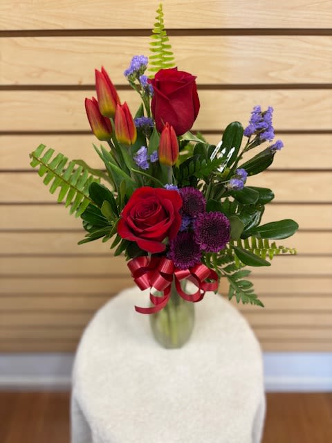 Wispy Dreams - Mixed cut arrangement with red roses, orange tulips, purple buttons, lavender statice, and mixed greens in a vase with a bow.