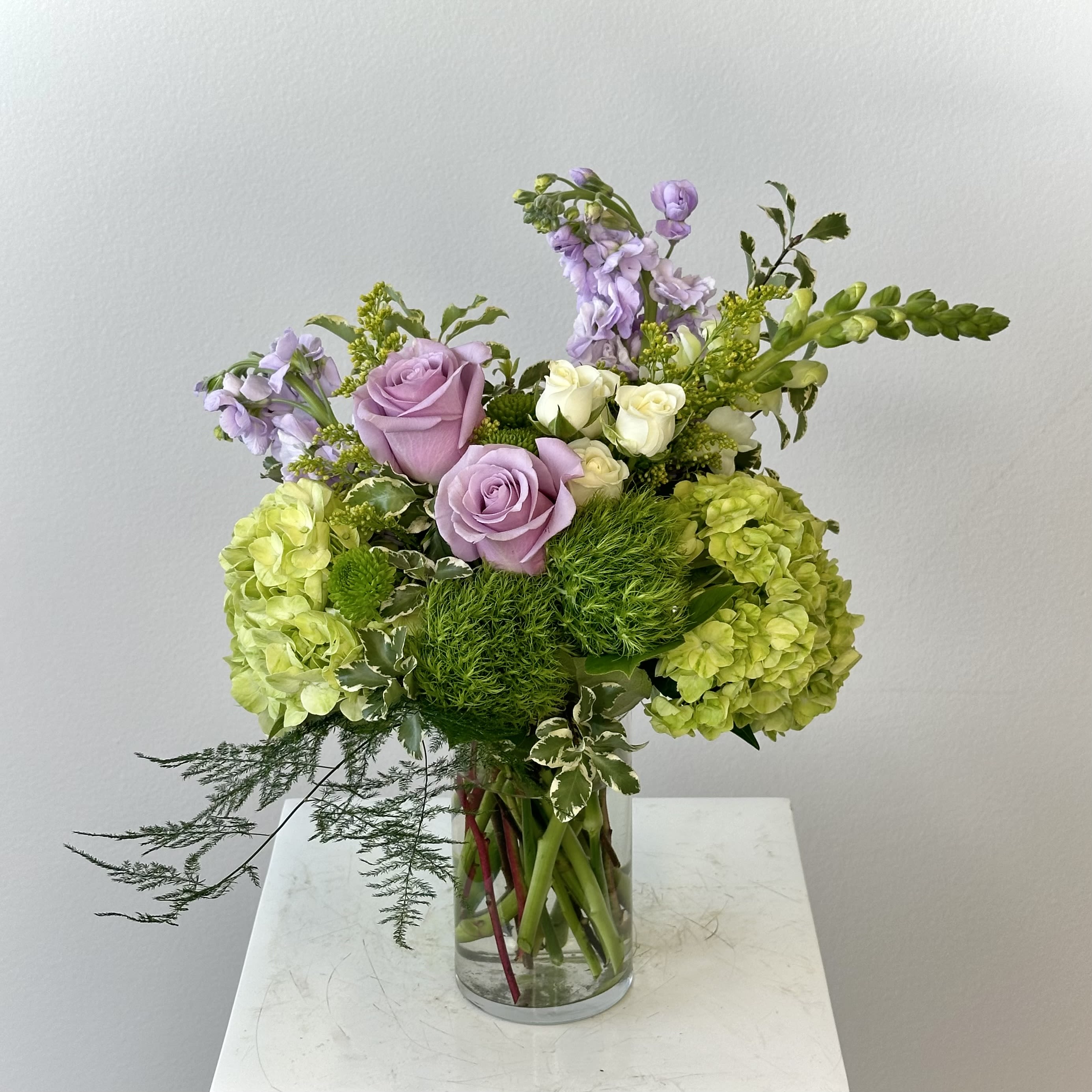 Lavender Matcha - Discover the serene elegance of the Lavender Matcha flower arrangement. This beautiful bouquet captures the calming essence of matcha with a sophisticated blend of soft lavenders, fresh greens, and crisp whites. Featuring luxurious lavender roses, lush green hydrangea, delicate white spray roses, fragrant lavender stock, charming dianthus balls, vibrant green button mums, and a tall white snapdragon, this design offers perfect harmony in color, texture, and fragrance. Lavender roses add romance and grace while the green hydrangea provides lush volume and natural energy. The white spray roses and snapdragon bring elegant contrast and purity. Fragrant lavender stock adds delightful height and scent, with green button mums and dianthus balls contributing unique texture. Perfect for birthdays, anniversaries, thank you gifts, or to create a peaceful home or office atmosphere. The Lavender Matcha’s unique purple and green palette makes it a modern alternative to traditional flower arrangements. Hand-arranged with the freshest premium flowers and delivered with care, this arrangement is designed to impress. Searching for lavender roses with green hydrangea? This is the ideal choice for a calming and stylish floral gift. Order the Lavender Matcha today and bring tranquil beauty into your life or someone special’s day.