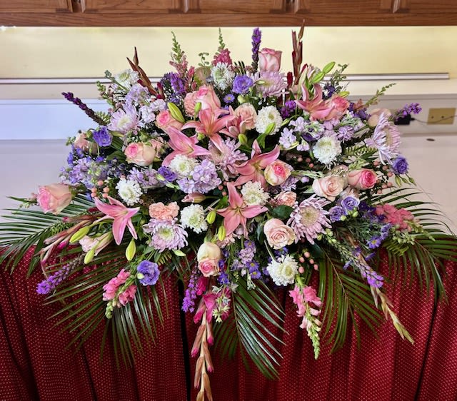 Grace in Bloom - Grace in Bloom is a lush, full-length casket spray designed to honor a beautiful life with softness and sophistication. Featuring elegant pink lilies, blush roses, lavender stock, delicate snapdragons, soft mums, and textured accent florals, this design is layered with gentle shades of pink, lavender, and cream. Flowing greenery and cascading palm fronds create graceful movement, giving the piece a natural garden feel while maintaining a timeless, refined presence.  Thoughtfully designed to offer comfort and beauty during a time of remembrance, Grace in Bloom serves as a heartfelt tribute — delicate, radiant, and full of love.