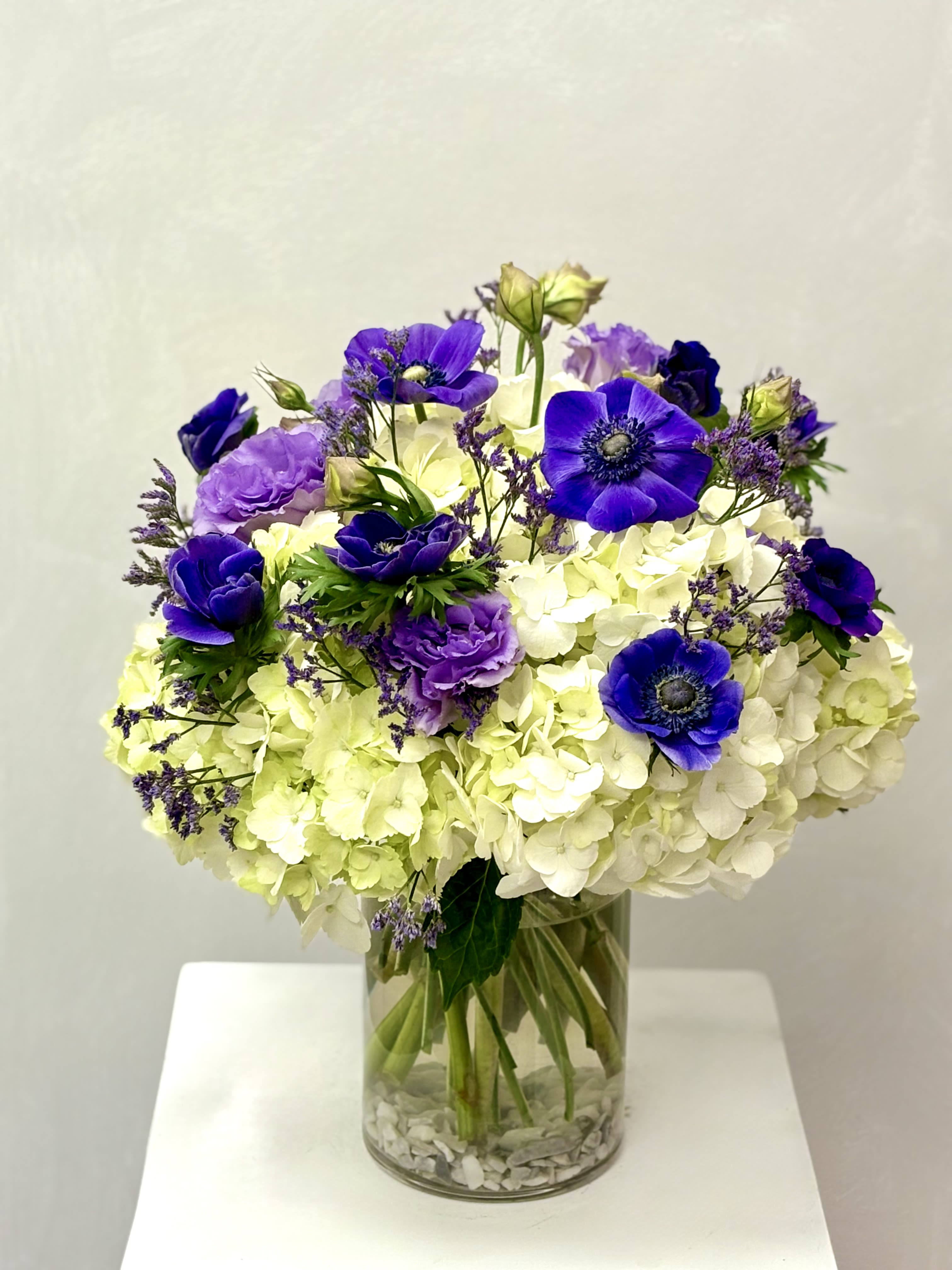 Royal Garden By VLM Flowers - A luxurious composition of creamy hydrangeas and bold violet anemones, highlighted by delicate purple blossoms. The rich color contrast and natural greenery create a fresh yet dramatic statement in a modern glass vase.