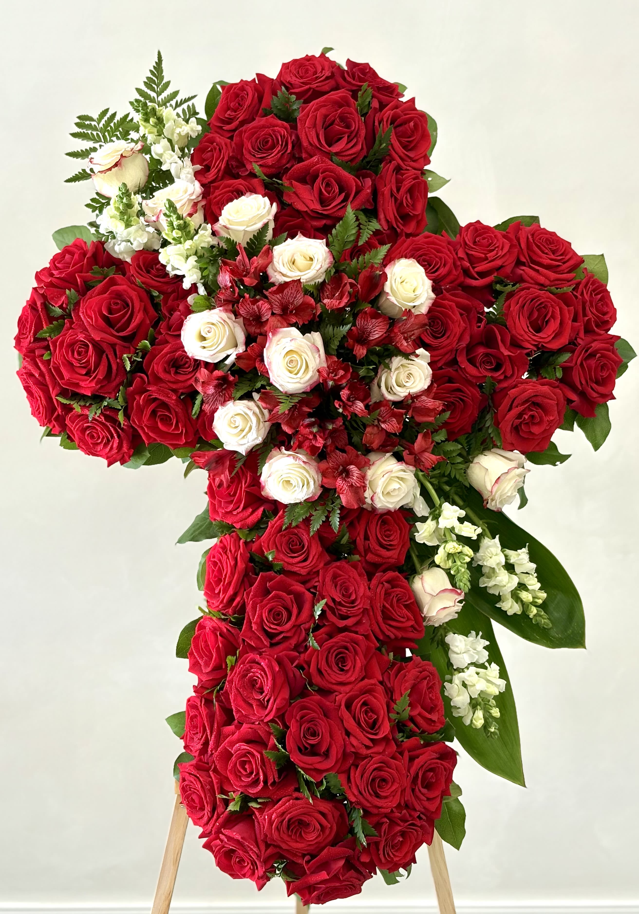 Sacred Love Cross By VLM Flowers - A beautifully crafted floral cross featuring lush red roses symbolizing deep love and devotion, accented with elegant white roses and soft white snapdragons for purity and peace. Clusters of red alstroemeria add texture and warmth, creating a rich, heartfelt expression of faith and remembrance. This striking tribute offers comfort and honors a loved one with grace, reverence, and timeless beauty.