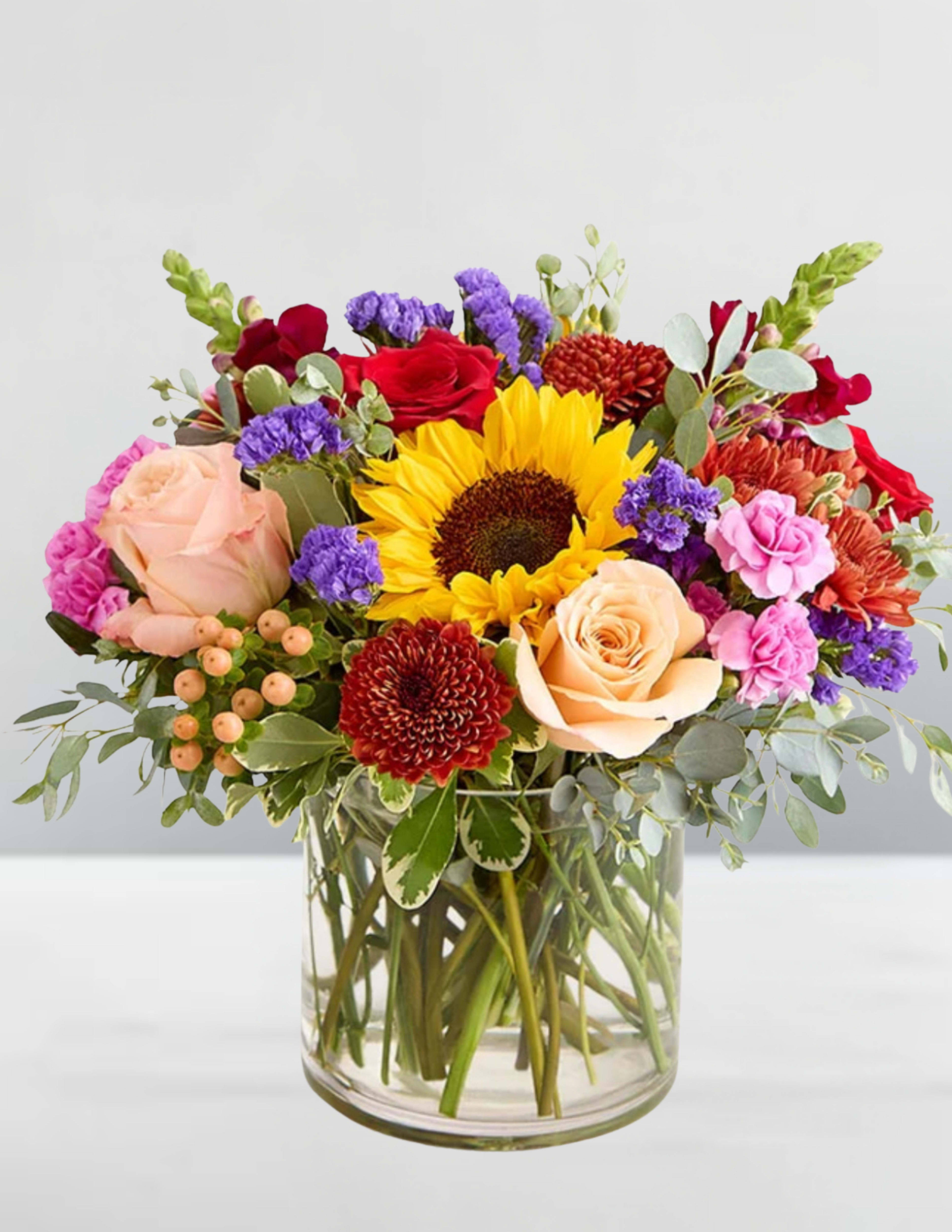 Letters from Autumn - Letters from Autumn A bouquet that feels like a handwritten note from the season itself—warm, wistful, and full of quiet beauty. At its heart, a golden sunflower beams like the last light of a fall afternoon, surrounded by crimson roses, peach and blush blooms, and the dusky purple of statice. Red chrysanthemums and pink carnations add texture and depth, while white berries and eucalyptus whisper of cooler days ahead.  This arrangement evokes the feeling of vintage postcards tucked in a drawer, the rustle of leaves underfoot, and the comfort of memories shared across time. Perfect for moments of reflection, gratitude, or simply to say “I’m thinking of you.