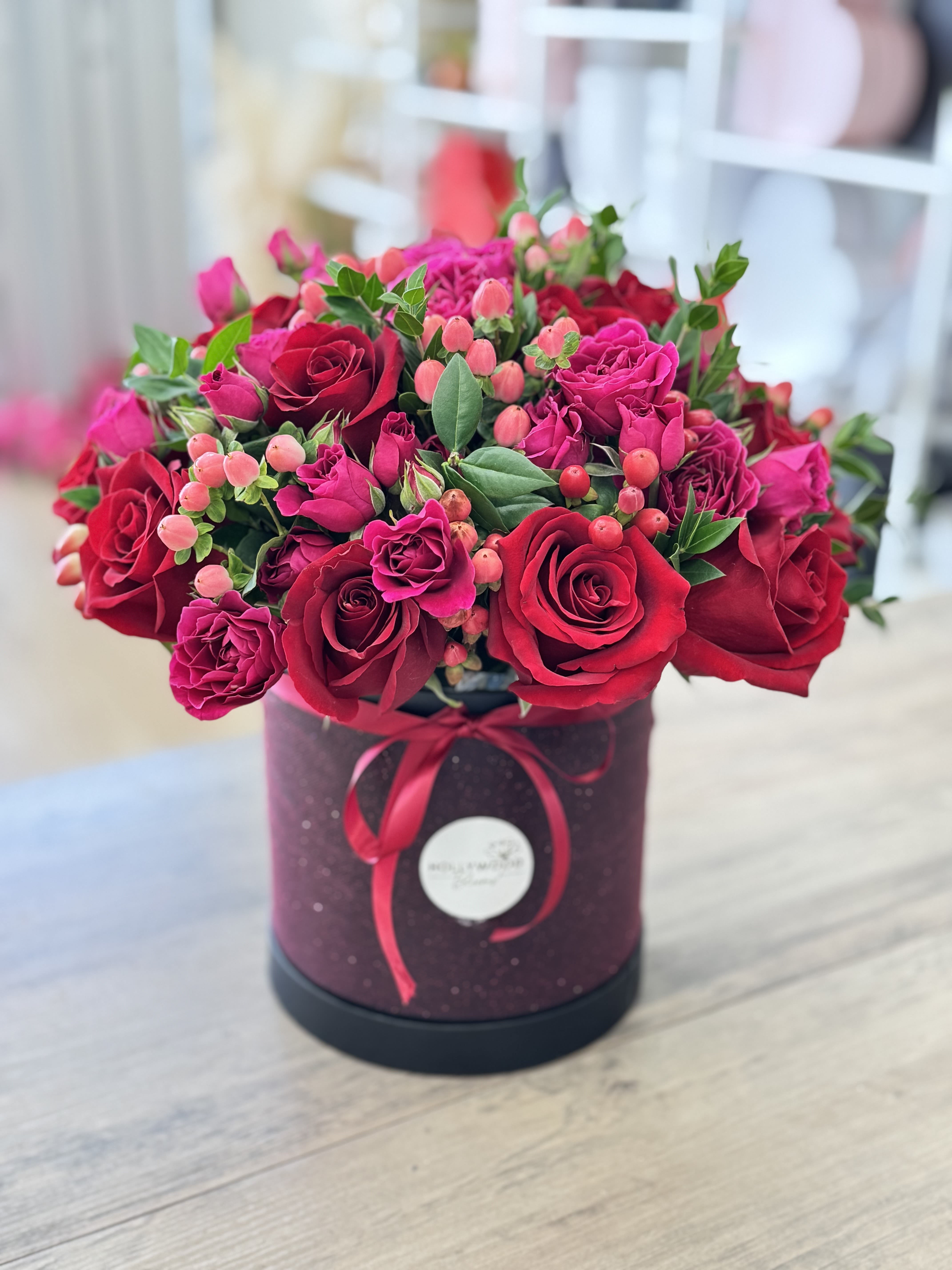 Red Velvet Flower Box - This luxurious arrangement is a bold celebration of romance, charm, and sophistication. Nestled inside a deep black hatbox with a sheer red bow, this design features a lush mix of velvety red roses, delicate pink spray roses, hypericum berries, and touches of silver eucalyptus for a soft metallic accent.  Rich in texture and full of passion, the bouquet’s color palette blends the timeless elegance of red with playful hints of soft pink, making it ideal for anniversaries, Valentine’s Day, romantic surprises, or elegant birthday gifts. The structured shape and compact design give it a refined look, while the vibrant blooms keep it expressive and full of life.  Whether you’re celebrating love or simply want to make a stunning statement, this arrangement is both luxurious and heartfelt — an unforgettable gift in a modern presentation.  Same-day flower delivery available in Los Angeles.
