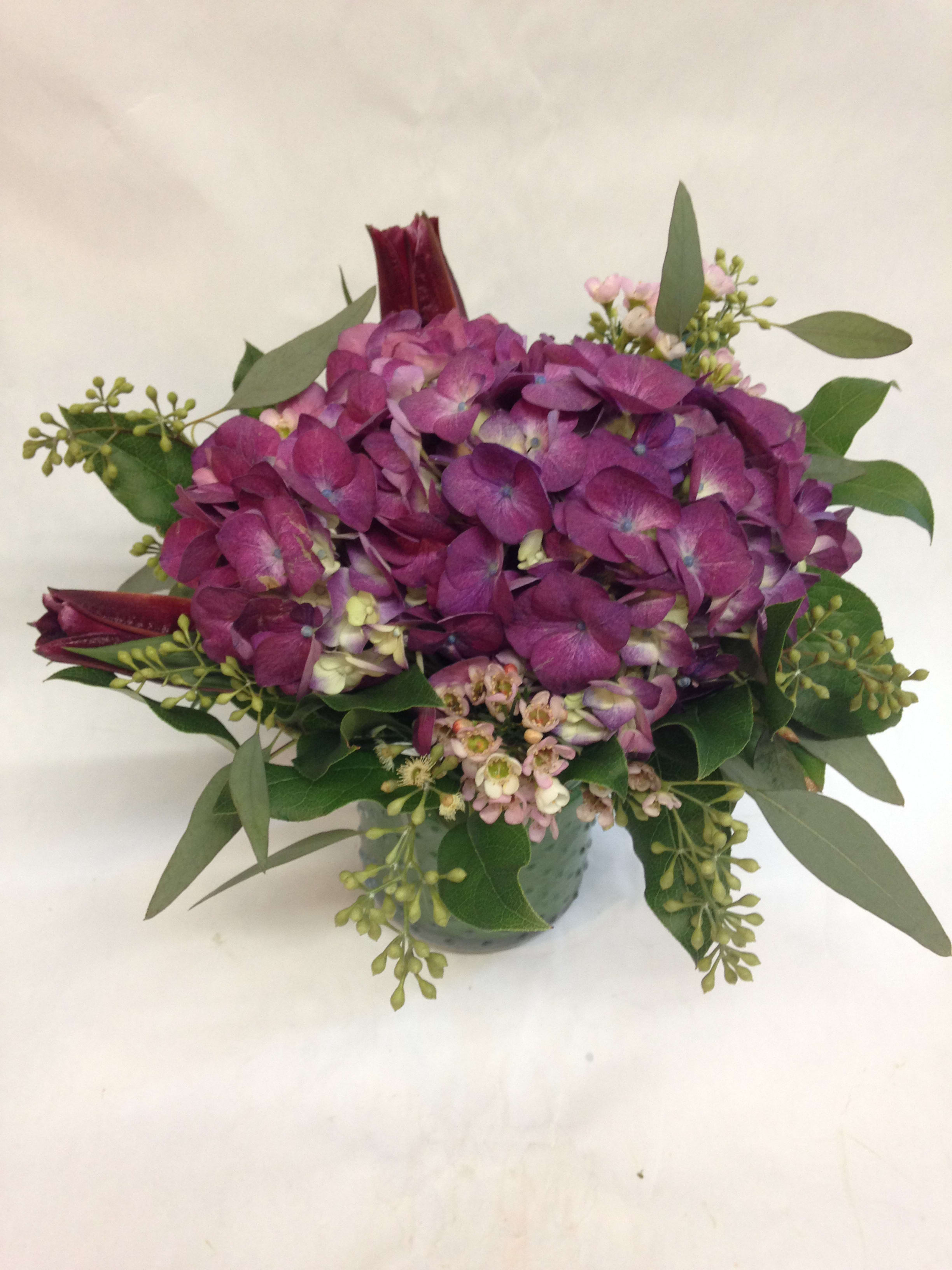 Simply Hydrangea - This centerpiece comes complete with a large hydrangea (color may be subject to change), burgundy lilies, wax flowers and seasonal greenery.  (Some flowers may be subject to change depending on the season. We will contact you with available flower substitutions; please contact us with specific questions if you are concerned with flower availability, thanks!) FEATURED ARRANGEMENT: $75.50 (REGULAR PRICE)