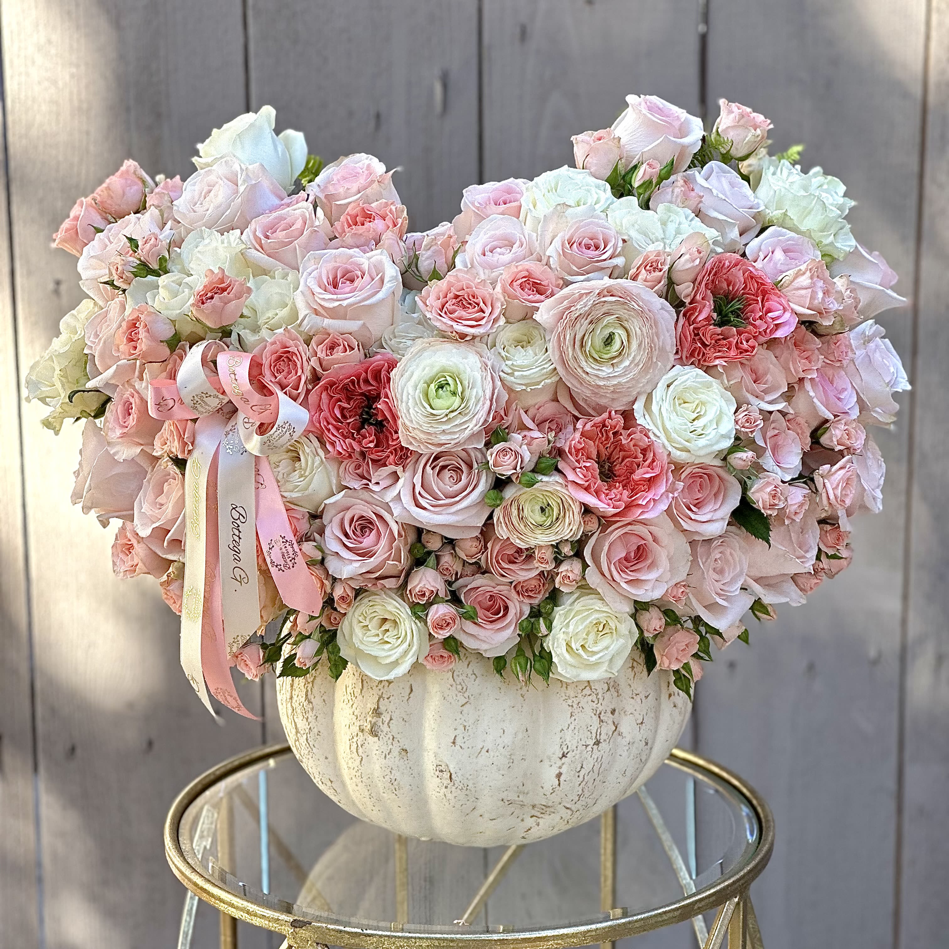 Blush Harvest - A white pumpkin serves as a charming vase for a floral arrangement featuring soft pink flowers. The pumpkin’s smooth, pale exterior contrasts beautifully with the vibrant blooms. Large pink roses, delicate peonies, and dainty ranunculus dominate the arrangement, adding texture and layers. Interspersed between the flowers are eucalyptus sprigs and wisps of baby’s breath, providing a touch of greenery and softness. The overall design has a whimsical, autumnal feel, blending the earthy warmth of fall with the romantic, fresh hues of pink. The arrangement is perfect as a centerpiece for a seasonal gathering.