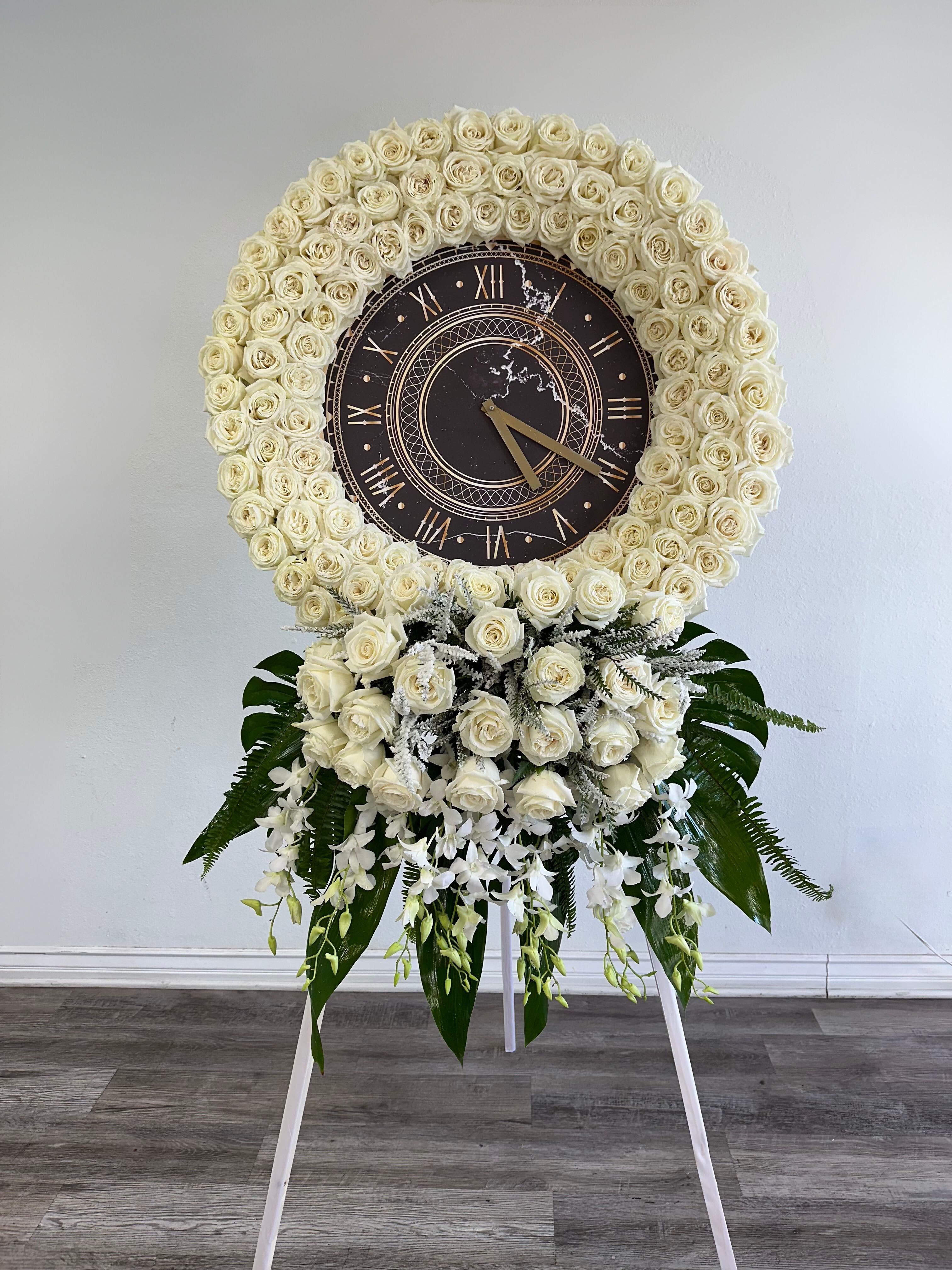 Timeless Tribute Wreath - The Timeless Tribute Wreath is a poignant and symbolic memorial, designed to honor a life that transcends time. At the center of this elegant standing wreath is a classic clock, representing the passage of time and the enduring legacy of a loved one. Surrounding the clock are pristine white roses, symbolizing purity, reverence, and eternal love. Lush greenery and delicate filler flowers complete the design, creating a peaceful and graceful tribute. This wreath serves as a beautiful reminder of cherished moments and the timeless impact a person leaves behind. Perfect for memorial services, it evokes a sense of peace and continuity.