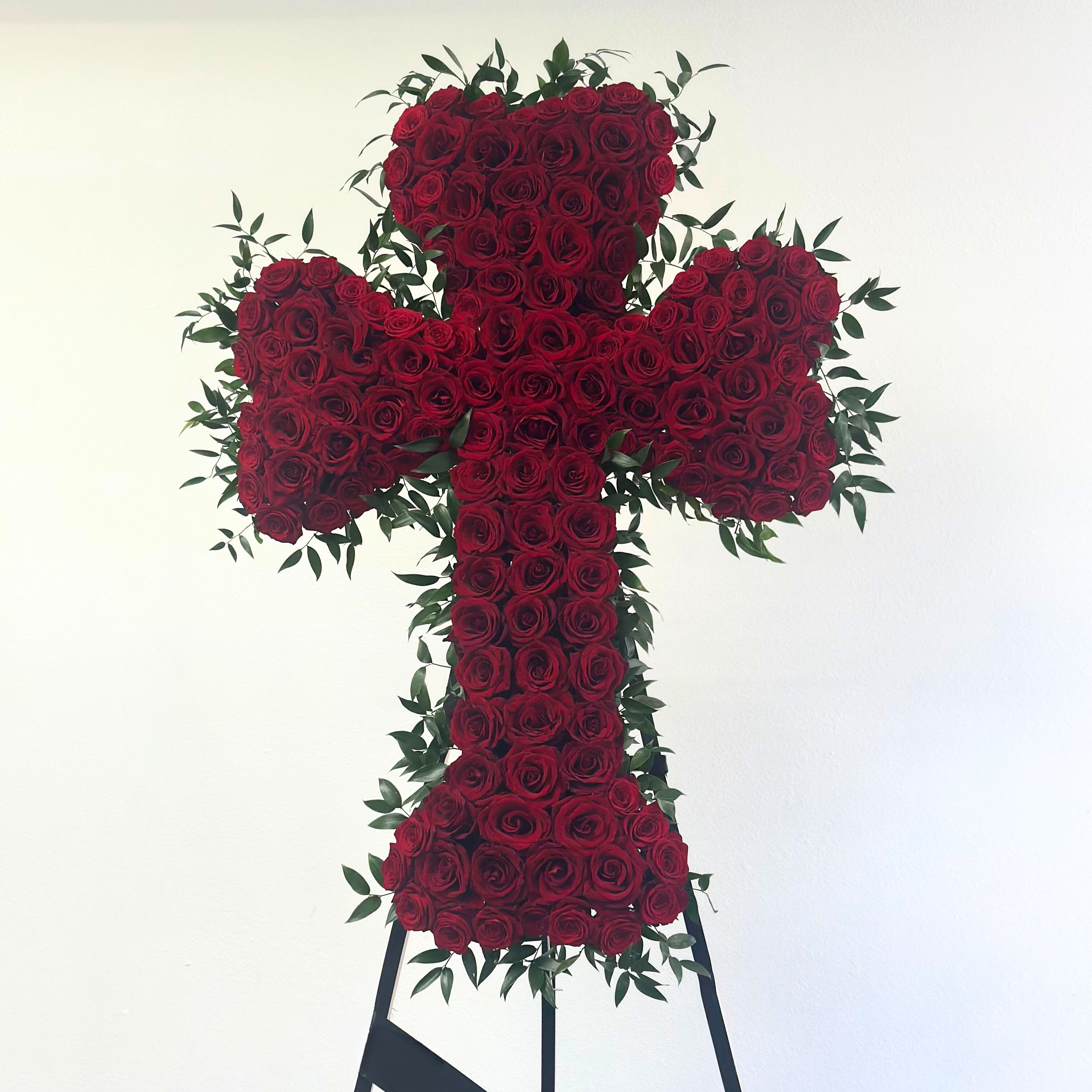 Blessed Memories Cross - Our Sympathy Standing Cross Wreath with Red Roses is a heartfelt and elegant tribute, perfect for honoring the memory of a loved one. This stunning arrangement features a gracefully designed cross shape adorned with lush greenery, white blooms for purity, and vibrant red roses symbolizing love and remembrance.