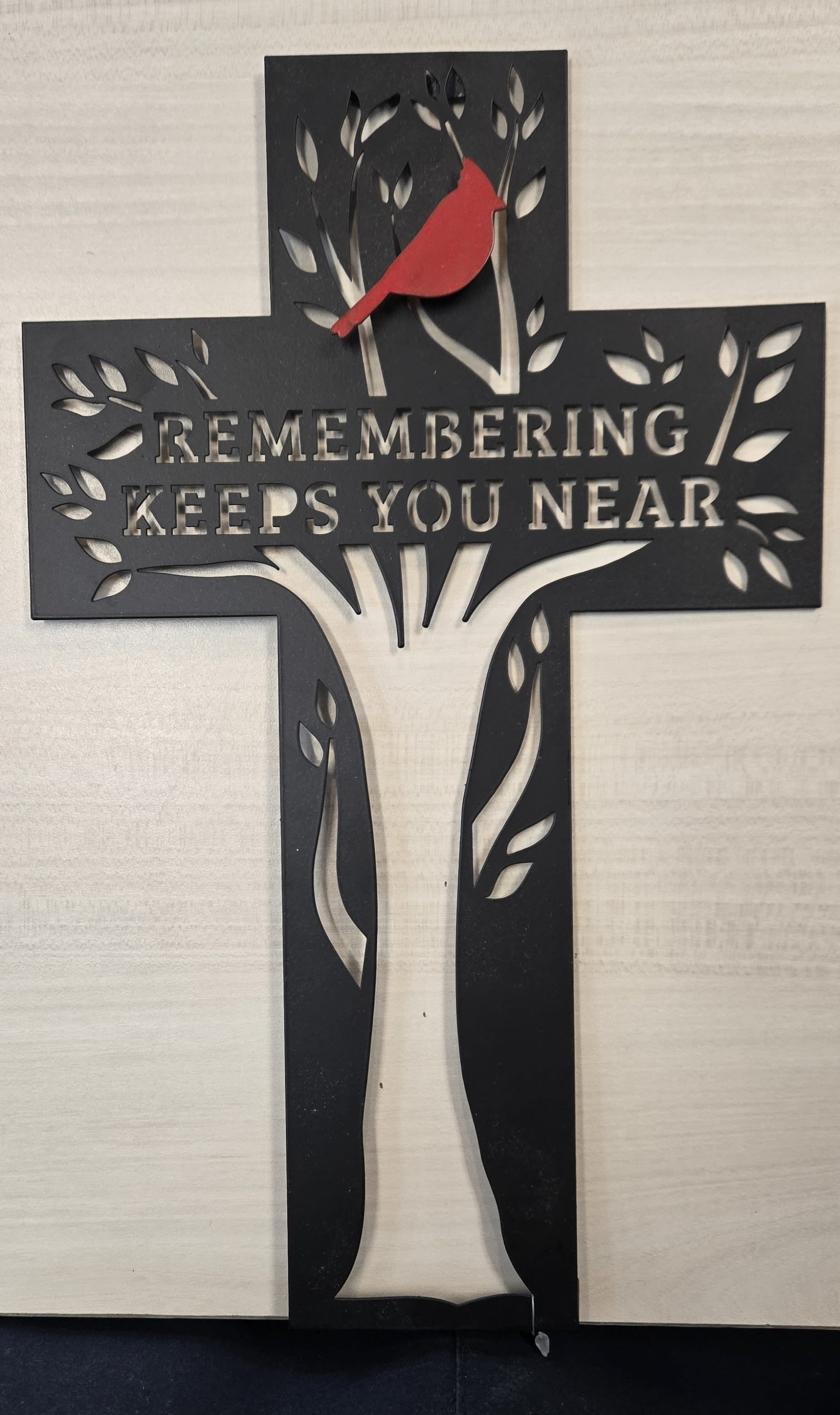 Cardinal of Remembrance - The photo shows a decorative wall cross made of dark metal. The cross features a stylized tree design cut into its surface, with branches and leaves spreading across the arms and trunk of the cross. Perched near the top is a bright red bird, which stands out vividly against the black metal. Across the center, the words “REMEMBERING KEEPS YOU NEAR” are cut into the metal, allowing the light-colored wall behind it to show through the lettering. The overall design combines faith, remembrance, and nature in a simple yet meaningful piece of art.
