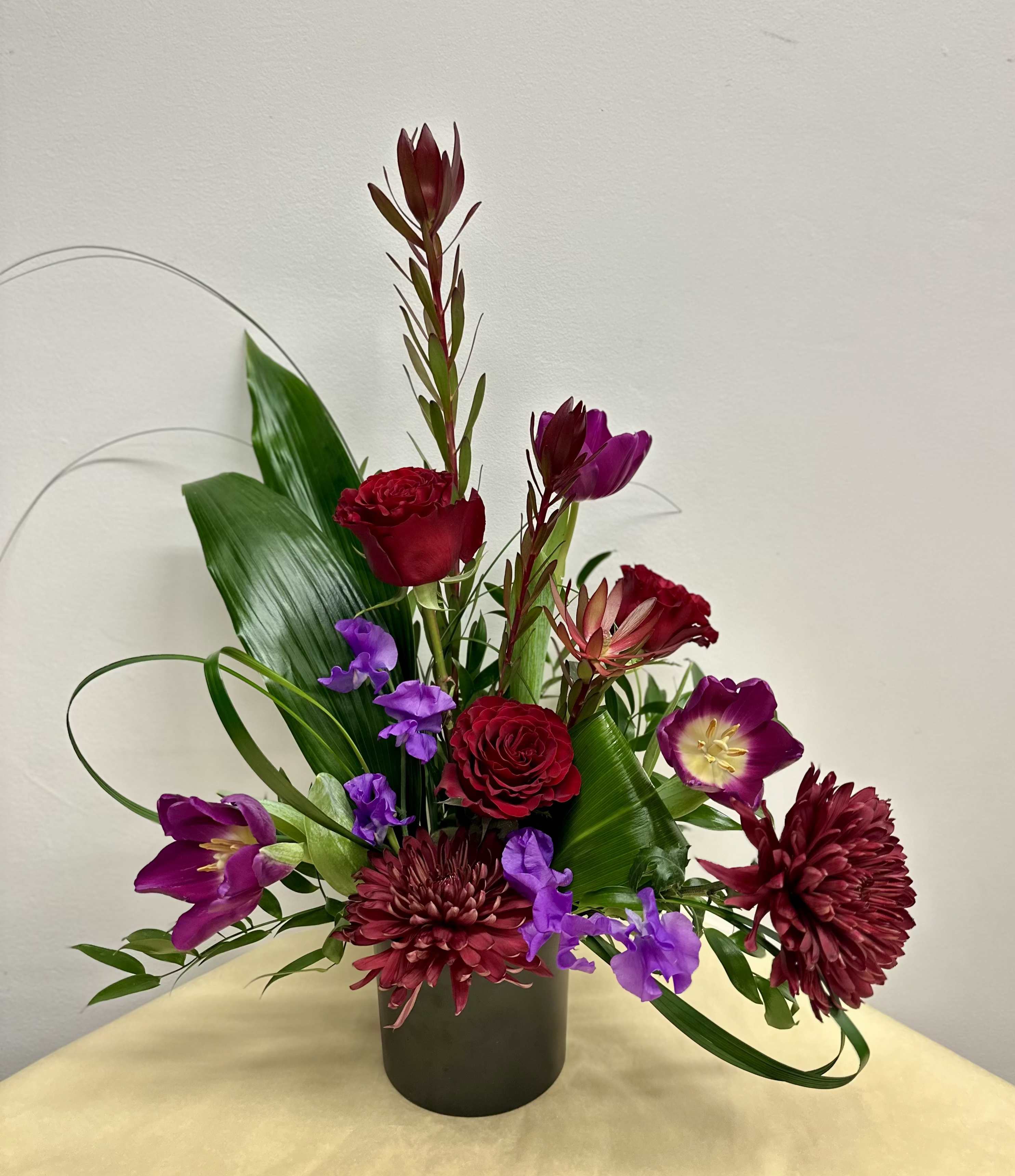 In The Mood -  A captivating arrangement that embraces deep, moody elegance with a bit of romance and intrigue.  This black glass container filled with rich red roses, velvety burgundy blooms, and dramatic purple flowers come together in a striking display.   