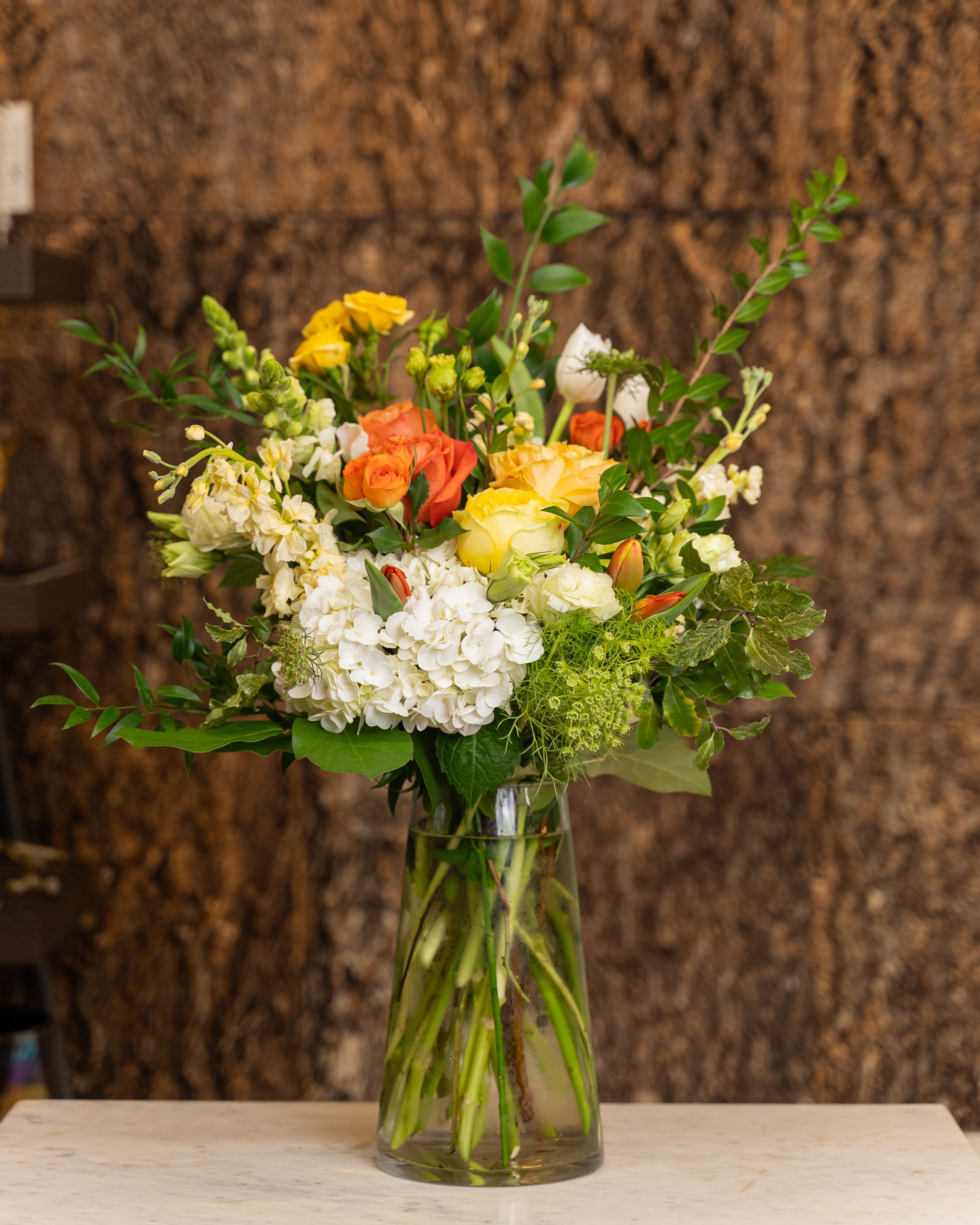 Flowers in Your Hair - Embrace the carefree, bohemian spirit of love with "Flowers in Your Hair". This whimsical arrangement captures the feeling of a sun-drenched afternoon and a romantic, playful connection. It features a joyful medley of sunny yellow roses, vibrant orange roses, crisp white hydrangeas, and delicate snapdragons, all accented with lush, flowing greenery. Presented in a clear glass vase, the airy design is reminiscent of a beautiful, natural crown of blossoms. It's a sweet, imaginative way to celebrate your unique relationship and the simple, natural beauty of being in love. 