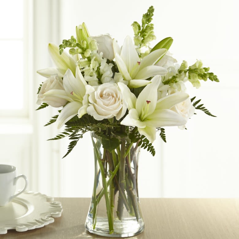 Eternal Friendship - An exuberance of bright and beautiful white blossoms provides an exquisite way to deliver your expressions of sympathy and comfort. This life affirming tribute combines white roses, snapdragons and Asiatic lilies accented by lush greens arranged in a clear glass 8" vase. An excellent choice for a wake, funeral service or for sending your condolences to the home of grieving family or friends.  Please Note: The bouquet pictured reflects our original design for this product. While we always try to follow the color palette, we may replace stems to deliver the freshest bouquet possible, and we may sometimes need to use a different vase.