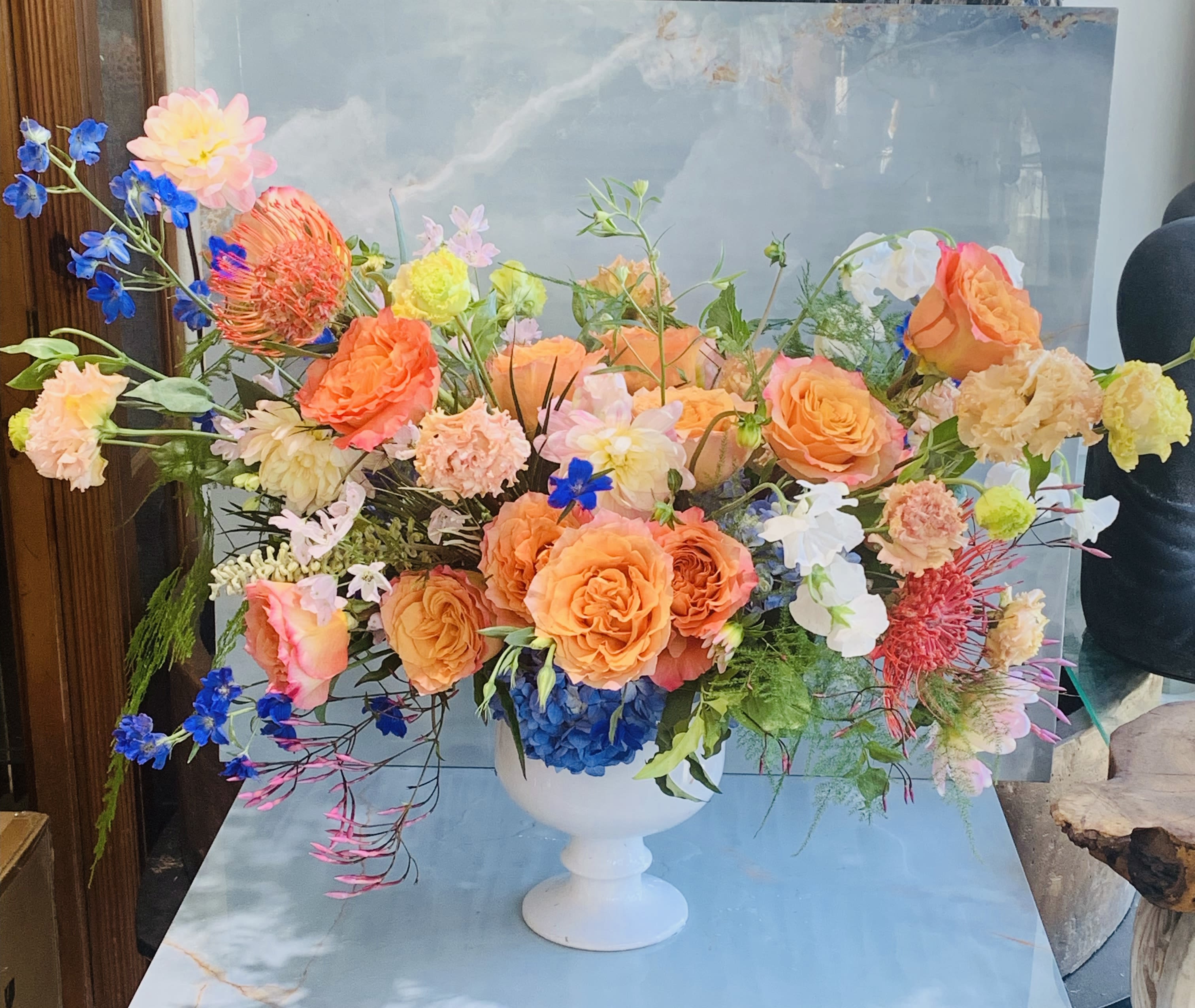 Peaches &amp; Pedals - This arrangement feels bright, garden-inspired, and joyful, with the coral and peach roses, blue delphinium, and playful textures giving it a lively spring garden feel. Accents of vivid blue and gentle pink create a lively contrast, while flowing greenery and whimsical stems add movement and a natural, freshly gathered feel. 