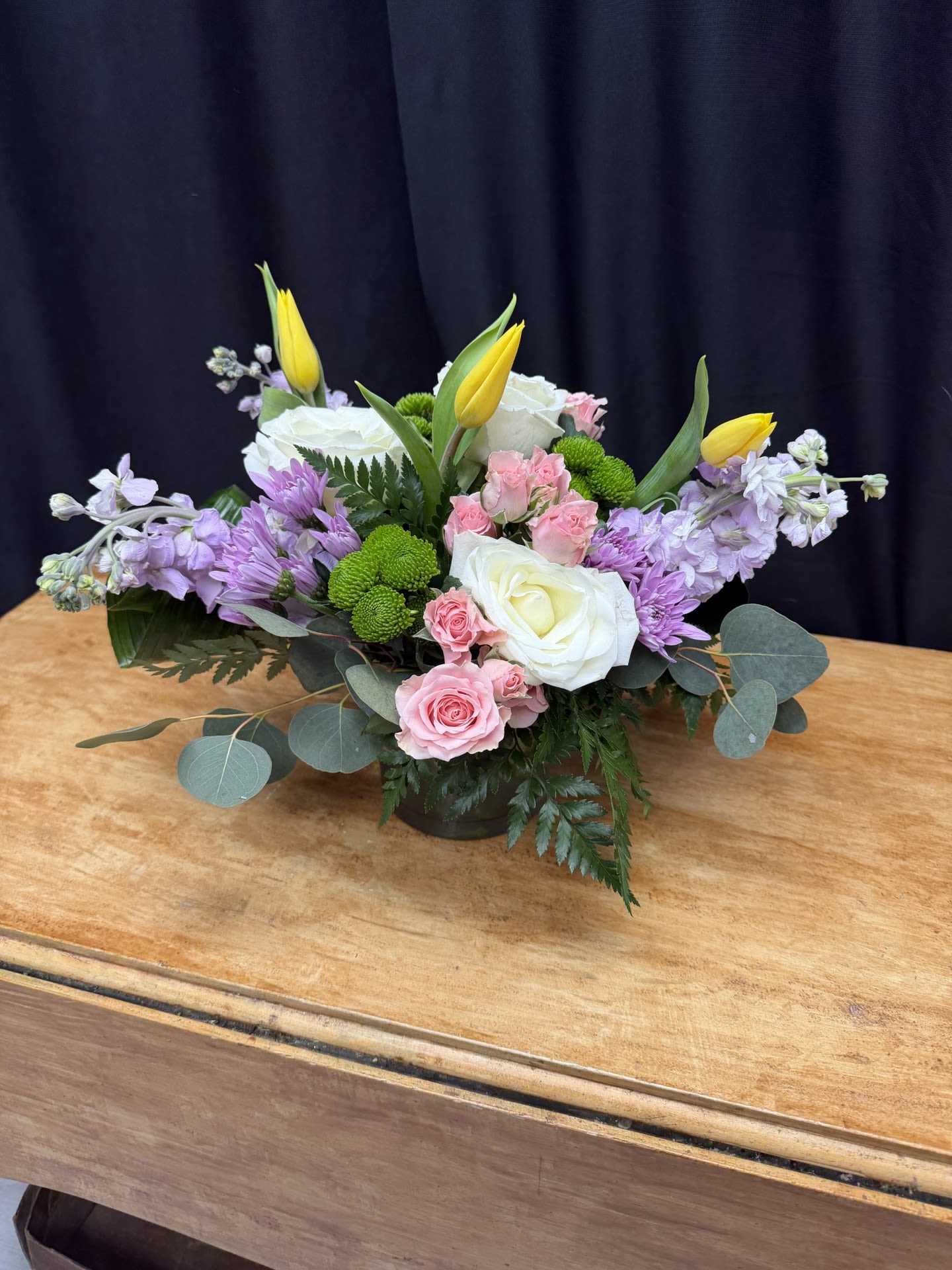 Spring Whisper - Spring Whisper is a graceful arrangement featuring soft pink roses, elegant white roses, cheerful yellow tulips, and delicate lavender stock. Accented with green button poms and fresh eucalyptus, the design brings together gentle spring colors in a naturally flowing style. This charming centerpiece adds a fresh, uplifting touch to celebrations, gatherings, or everyday moments. 