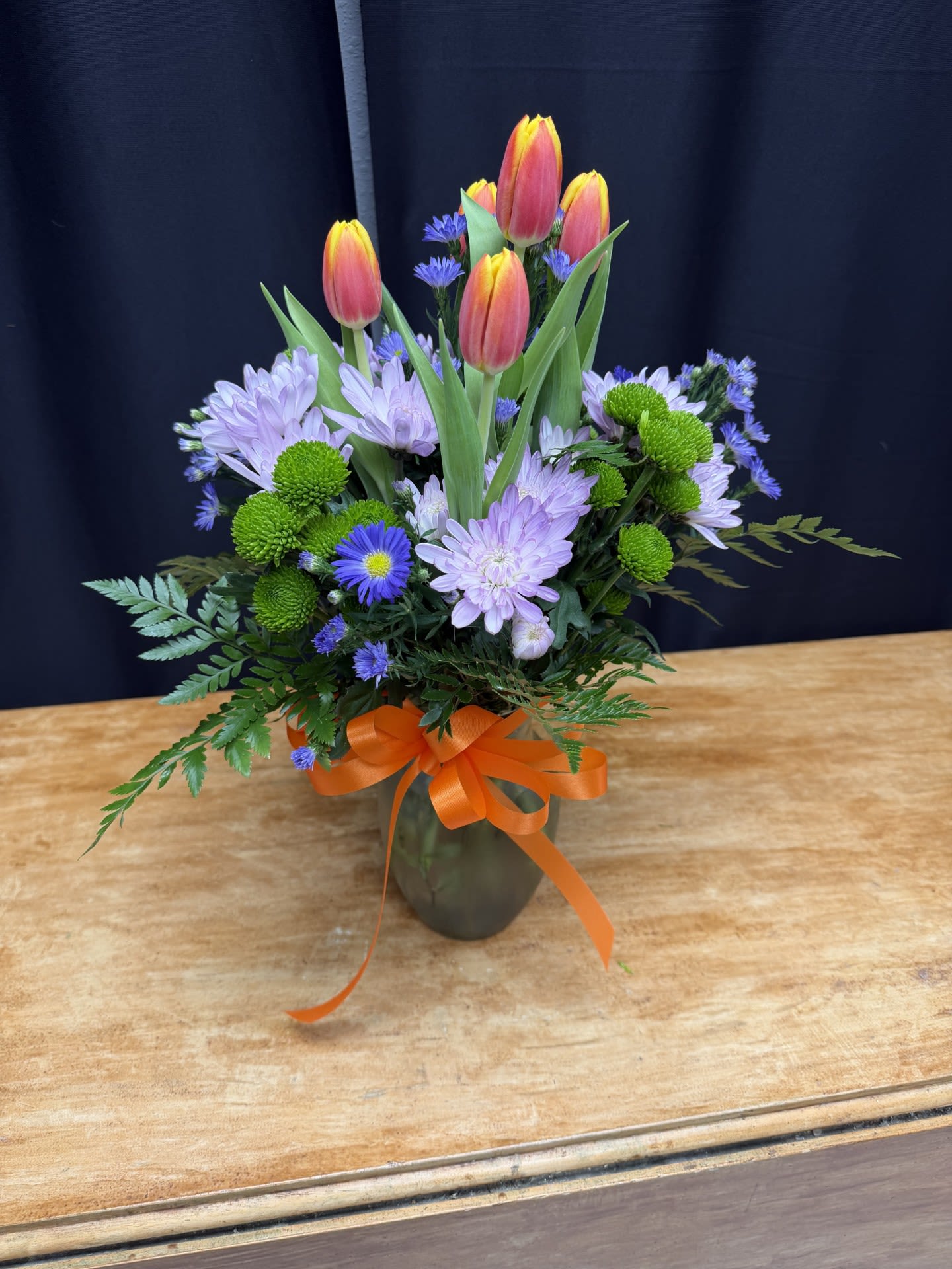 Spring Sunrise - Spring Sunrise features vibrant sunset tulips rising above soft lavender chrysanthemums, bright green button poms, and delicate purple asters. Fresh greenery adds texture and movement, while a cheerful ribbon completes the garden-inspired design. This lively arrangement captures the fresh colors and joyful energy of spring, perfect for brightening any room. 