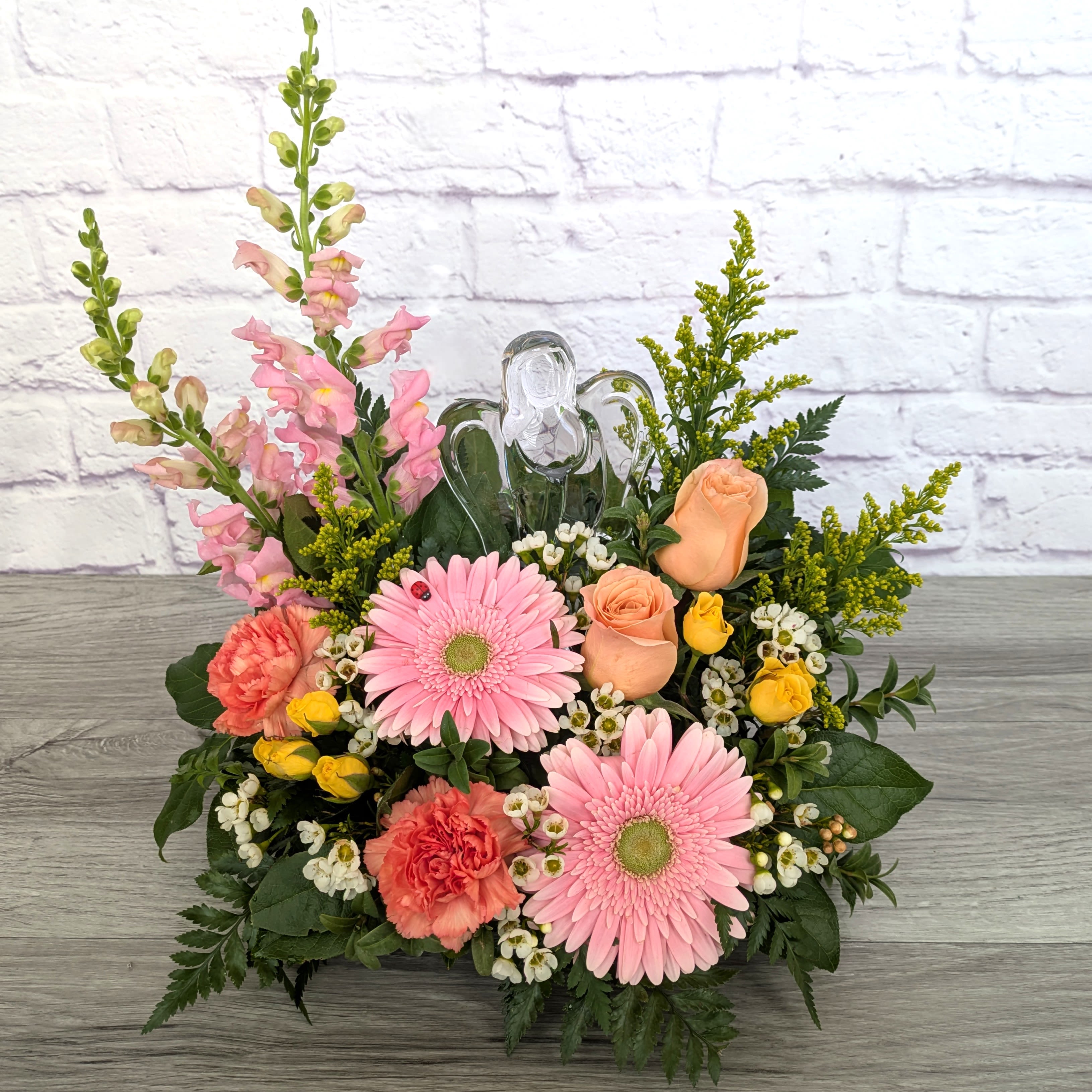 Angelic Embrace - Angelic Embrace is A Ladybug Floral exclusive bouquet. This bouquet features snapdragons, gerbera daisies, roses, spray roses, solidago, and wax flower. All lovingly designed in an oasis dish with a 6” crystal angel.  Orientation: One Sided 16” Tall 14” Wide  