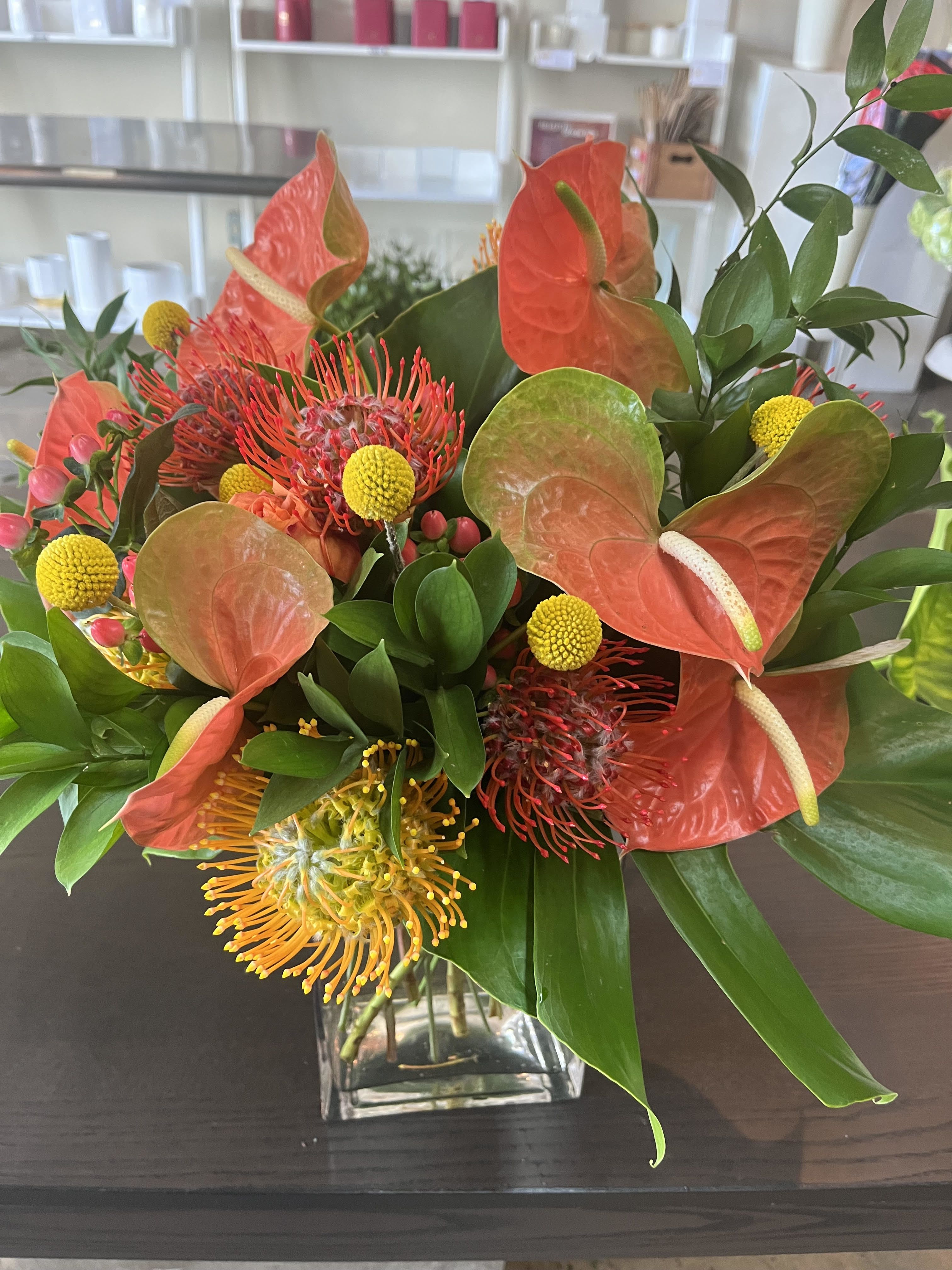 Flora Tropical - low arrangement in a square crystal vase with beautiful Anthuriums, pincushion proteas, Hypericum, Craspedia, Monstera, Israeli Ruscus, Italian Ruscus.   Great as a centerpiece for a dinner 0 or as a gift!   