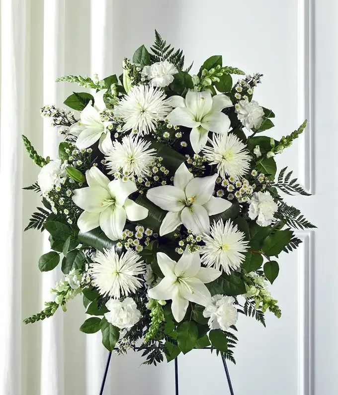 Graceful White Tribute – Standing Spr - A timeless and elegant white standing spray designed to honor and celebrate the life of a loved one.  This arrangement features a serene palette of fresh white flowers including lilies, chrysanthemums, carnations, and delicate chamomile accents, surrounded by lush greenery. The design creates a soft, peaceful tribute suitable for funerals, memorial services, and celebrations of life.  The arrangement is presented on a standing easel, making it ideal for display beside the casket, memorial table, or photo tribute.  Each piece is handcrafted with care and may vary slightly depending on seasonal flower availability while maintaining the same overall style and color palette.  Includes easel stand.