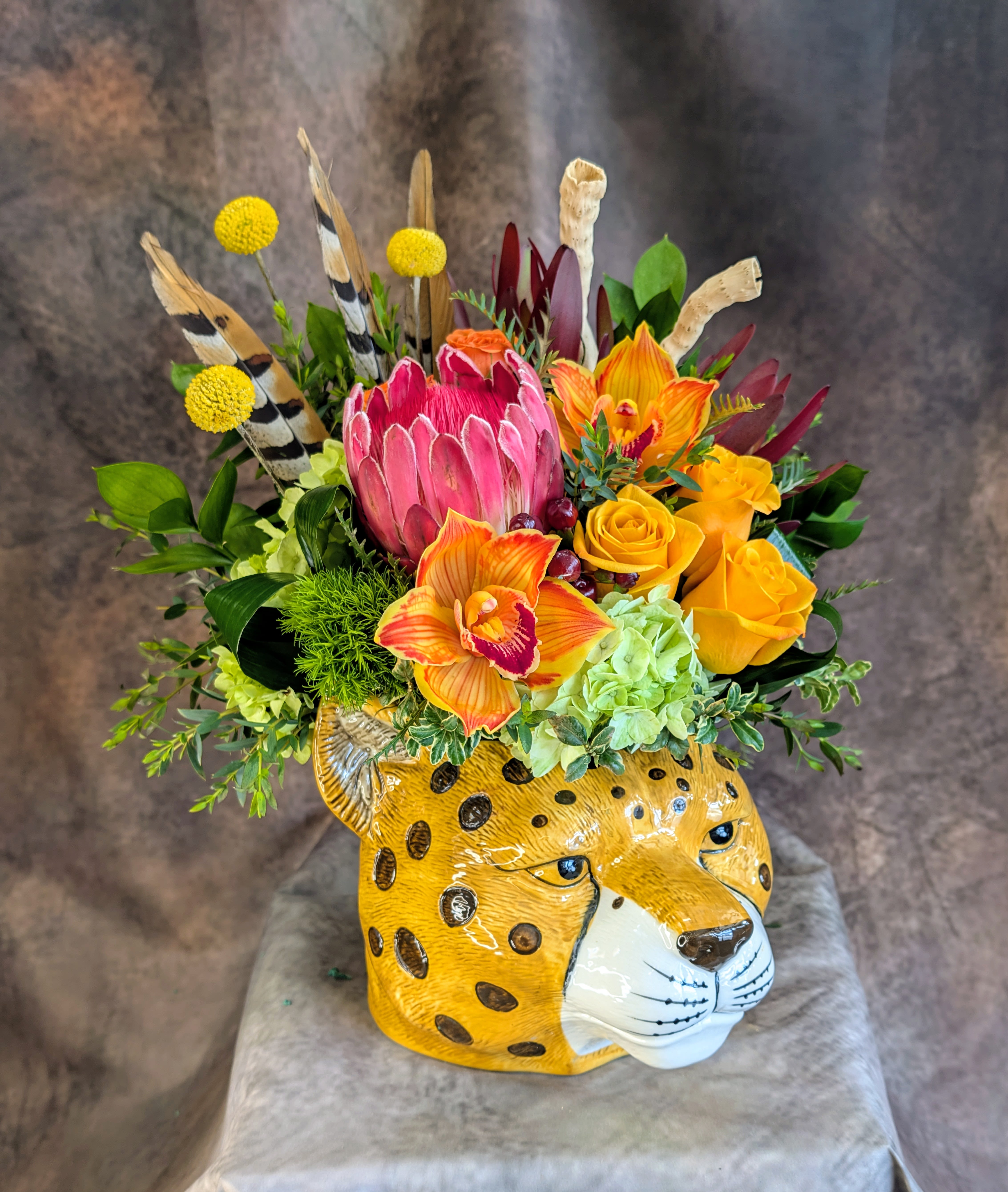 Untamed - Bold. Wild. Unapologetically beautiful.  Untamed is a striking, safari-inspired floral design created for those who love florals with personality. Designed in a sculptural ceramic leopard vessel, this arrangement embodies the spirit of adventure with vibrant color, dramatic texture, and unexpected elements that feel both luxurious and wildly natural.  At the heart of the design sits a magnificent pink protea, surrounded by glowing golden roses and fiery orange cymbidium orchids that radiate warmth and energy. Soft green hydrangea, lush seasonal foliage, and textured botanicals add depth and movement, while craspedia billy balls, natural feathers, and Gobi wood  accents create an untamed, organic silhouette.  The artistic cheetah vessel transforms this piece into a true statement arrangement—perfect for those who appreciate bold décor, modern botanical design, and pieces that spark conversation.  Handcrafted by Windmill Floral Expressions, a family-owned Las Vegas and Henderson florist, Untamed is ideal for birthdays, celebrations, luxury gifting, or anyone who embraces a little wild beauty in their space.  Each arrangement is thoughtfully designed with premium seasonal blooms and artistic flair, making every piece one-of-a-kind.