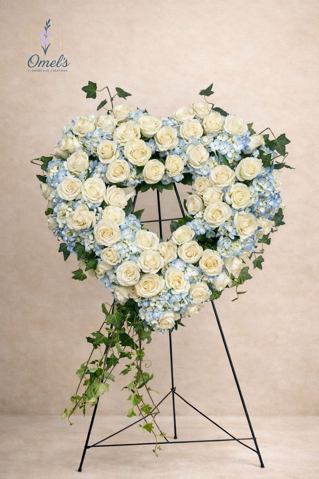 Heavenly Heart Tribute - A graceful expression of everlasting love, the Heavenly Heart Tribute is designed in a classic open-heart shape and beautifully arranged on a standing easel. This elegant sympathy piece features lush white roses nestled among soft blue hydrangeas, accented with trailing ivy for a natural, flowing finish.  The serene combination of white and soft blue symbolizes peace, remembrance, and heartfelt devotion — making it a meaningful choice for funeral services, memorials, or celebration of life ceremonies.  Handcrafted with care and compassion, this tribute offers a beautiful way to honor and celebrate a life well lived.