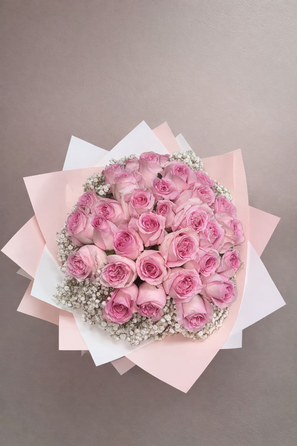 25 Pink Roses Wrap - A timeless expression of love and grace, this stunning bouquet features a lush arrangement of soft pink roses beautifully nestled among delicate white baby’s breath. Wrapped in layered blush and white premium paper, the design creates a romantic, cloud-like presentation that feels both modern and classic.  Each rose is hand-selected for its full bloom and gentle pink hue, symbolizing admiration, sweetness, and heartfelt affection. Perfect for Valentine’s Day, anniversaries, birthdays, or simply to brighten someone’s day, this bouquet makes a sophisticated statement without saying a word.  Soft, feminine, and effortlessly elegant — a bouquet that turns every moment into something unforgettable. 
