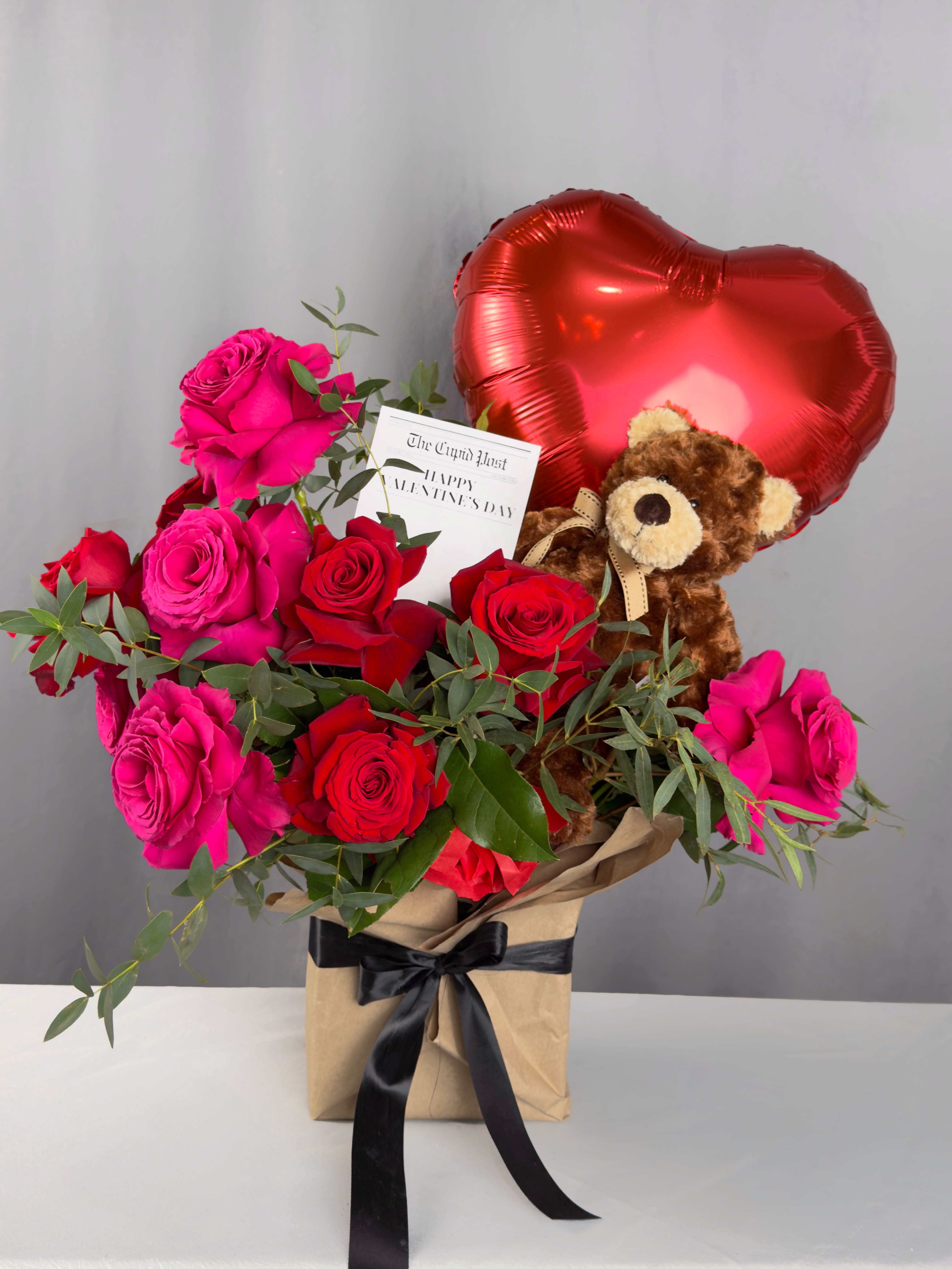Teddy Love Story - Romantic floral arrangement featuring 10 fresh roses, accented with seasonal fresh greens, and wrapped in natural kraft paper for a clean, elegant look. Includes a plush teddy bear and a Mylar balloon, making it the perfect gift to sweetly celebrate any special occasion.