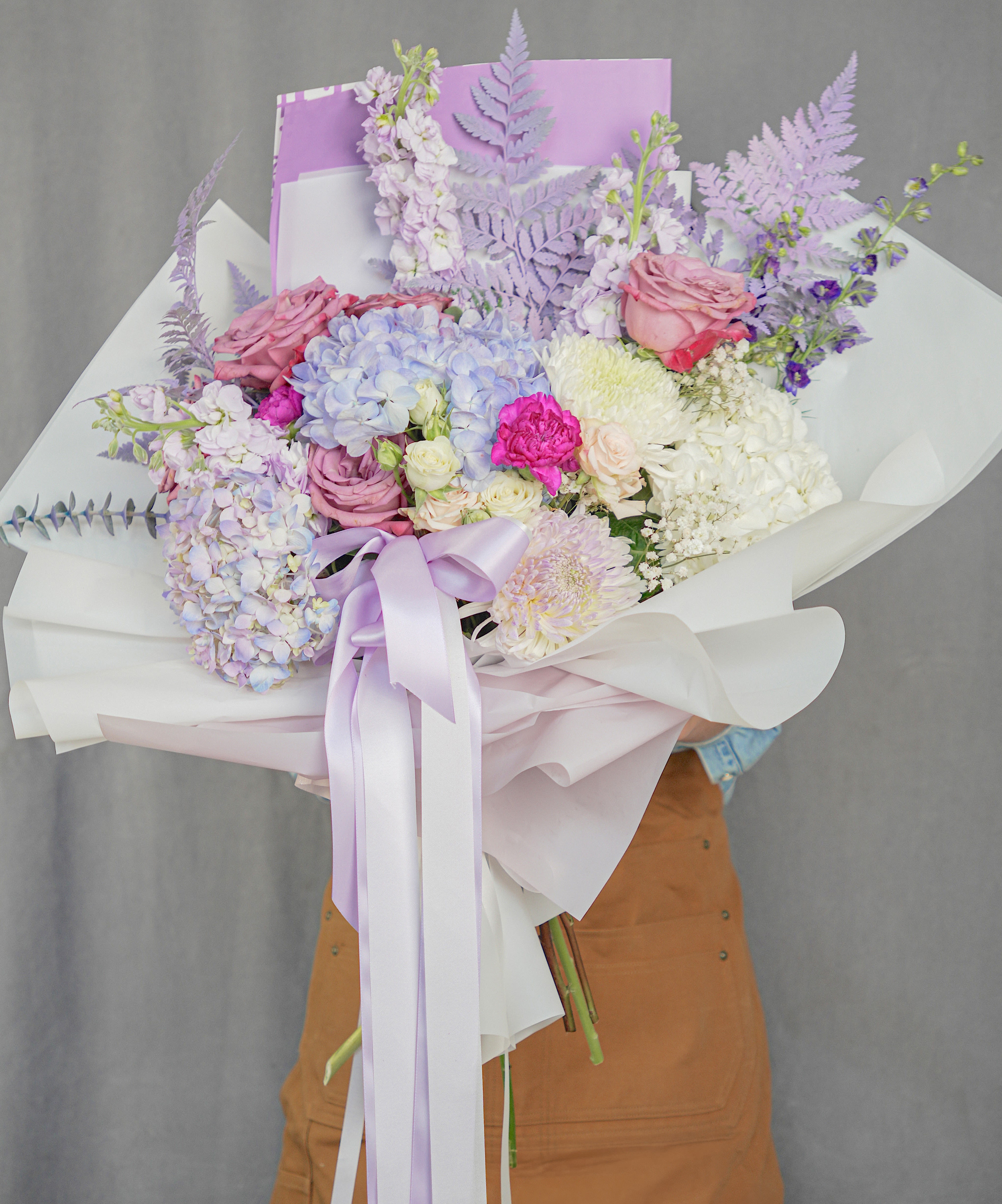 LAVENDER DREAM  - This elegant bouquet is wrapped in premium Korean-style paper and designed with seasonal flowers in soft lavender and blush tones. The arrangement may include a selection of premium blooms such as roses, hydrangeas, stock, snapdragons, leather leaf, eucalyptus, spray roses, carnations, mums, baby’s breath, and hand-painted lavender foliage. Finished with soft fabric ribbons in lavender for a delicate and romantic touch. Please note that flower varieties may vary depending on seasonal availability, while maintaining the same color palette and overall style.*